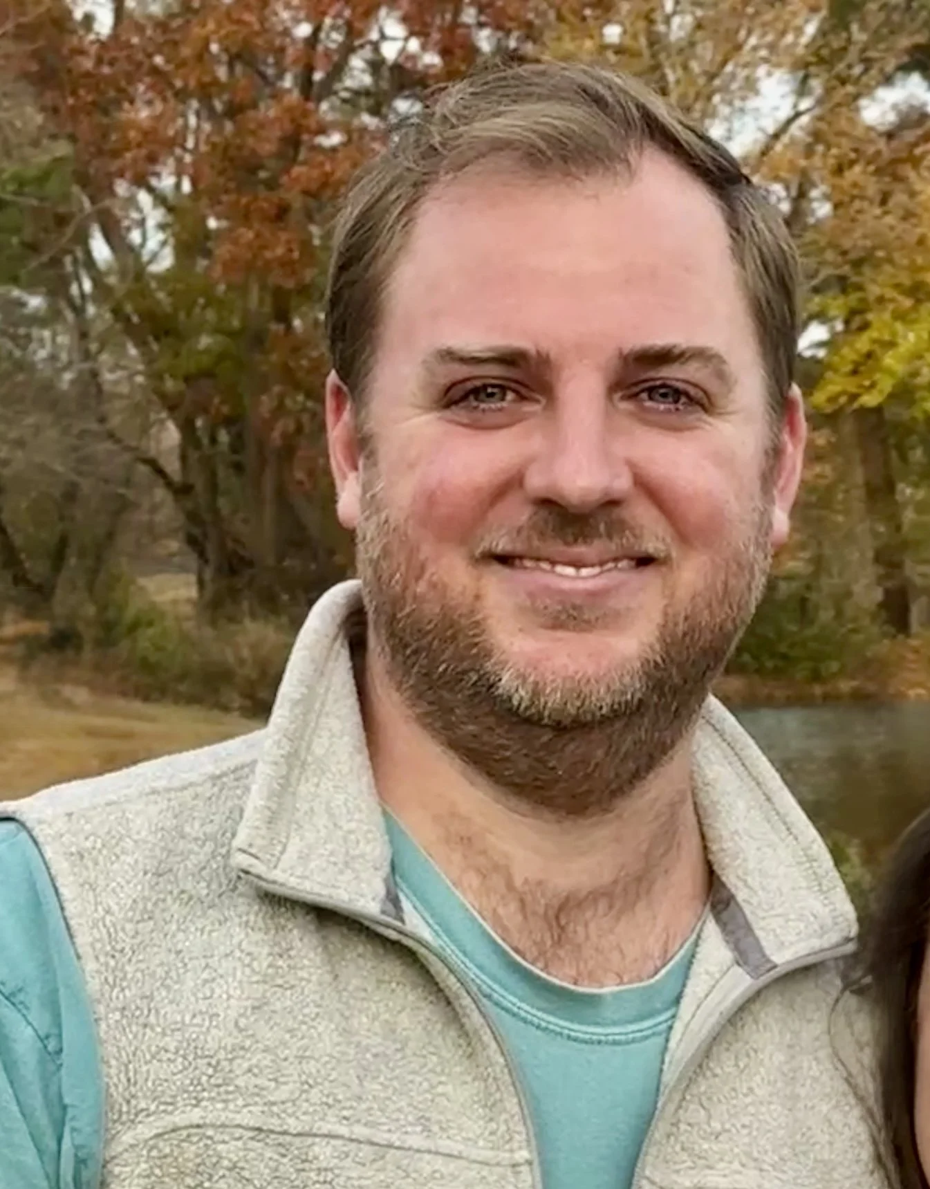 A close-up of a smiling man with brown hair and a beard, wearing a light gray vest over a teal shirt, outdoors during fall with trees whose leaves are changing color in the background.