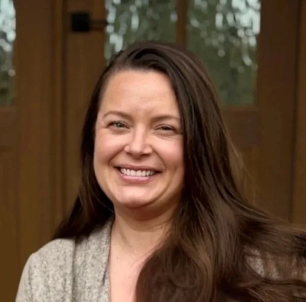 A woman with long brown hair smiling in front of a wooden background with windows.