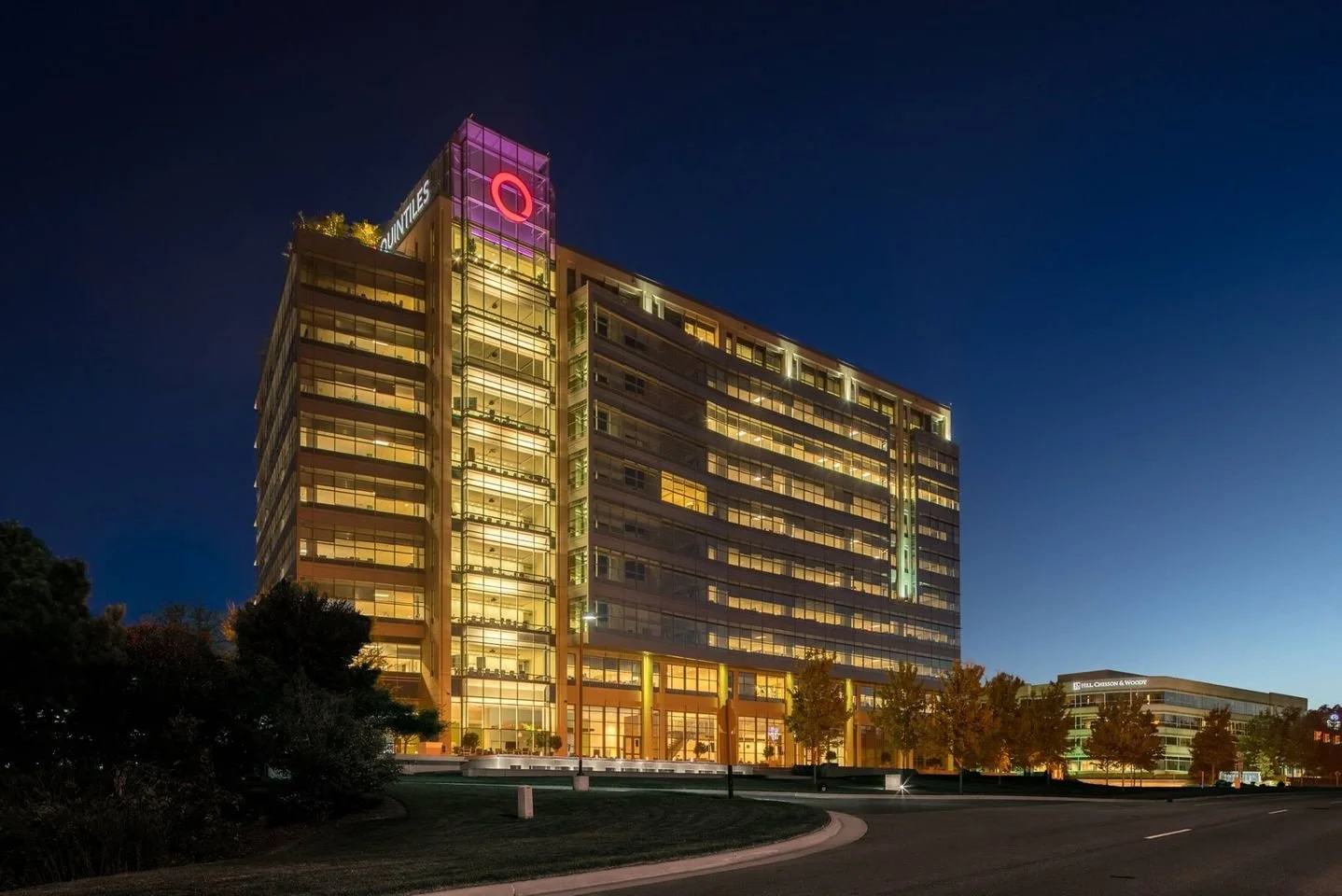 Nighttime view of a lit-up modern office building with glass windows, trees in front, and a dark sky in the background.