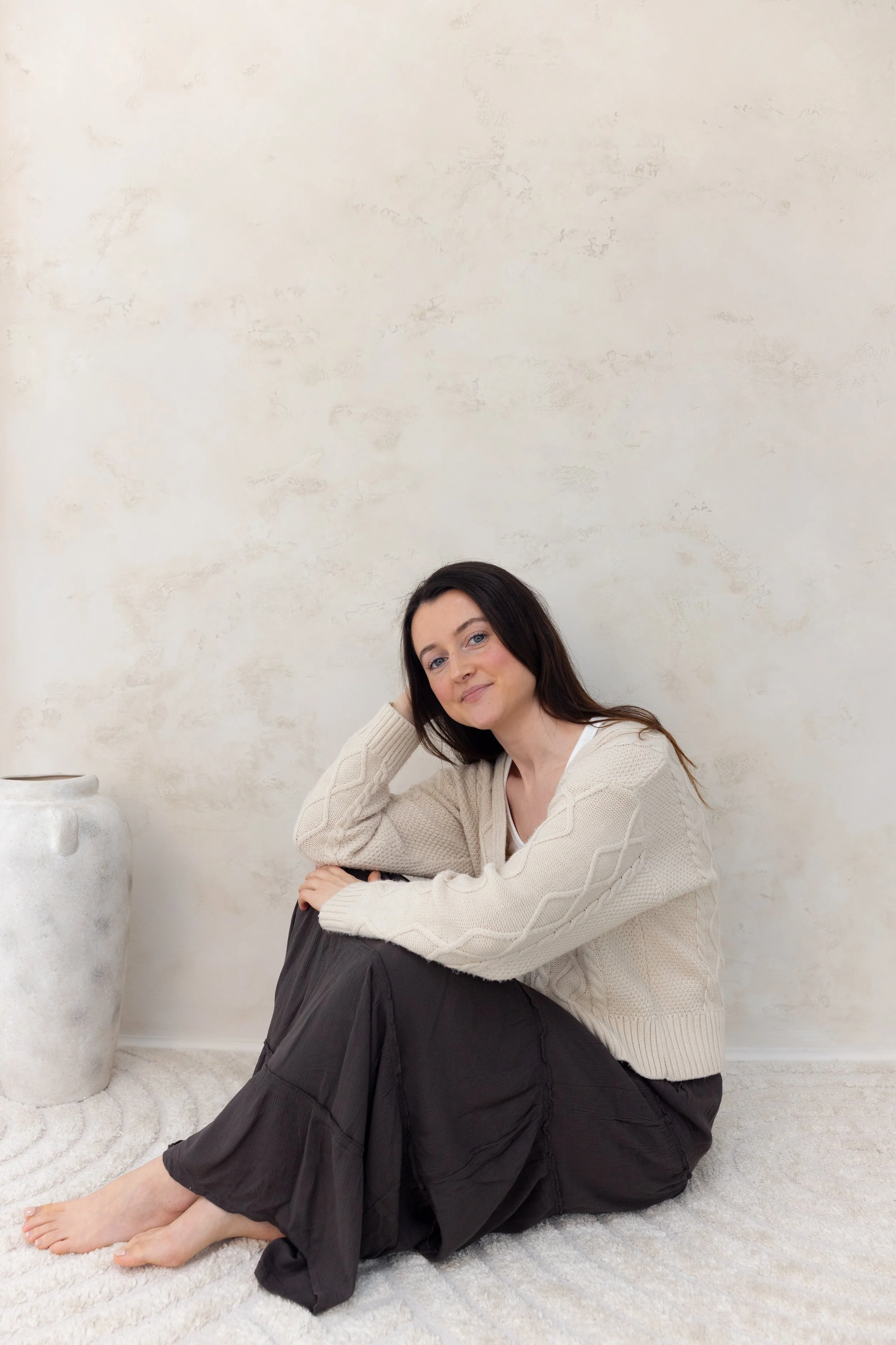 A woman with long dark hair sitting on a white rug, resting her arm on her knees. She is wearing a beige knitted sweater and a black skirt. There is a large white ceramic vase next to her on the left. The background is a plain, textured white wall.