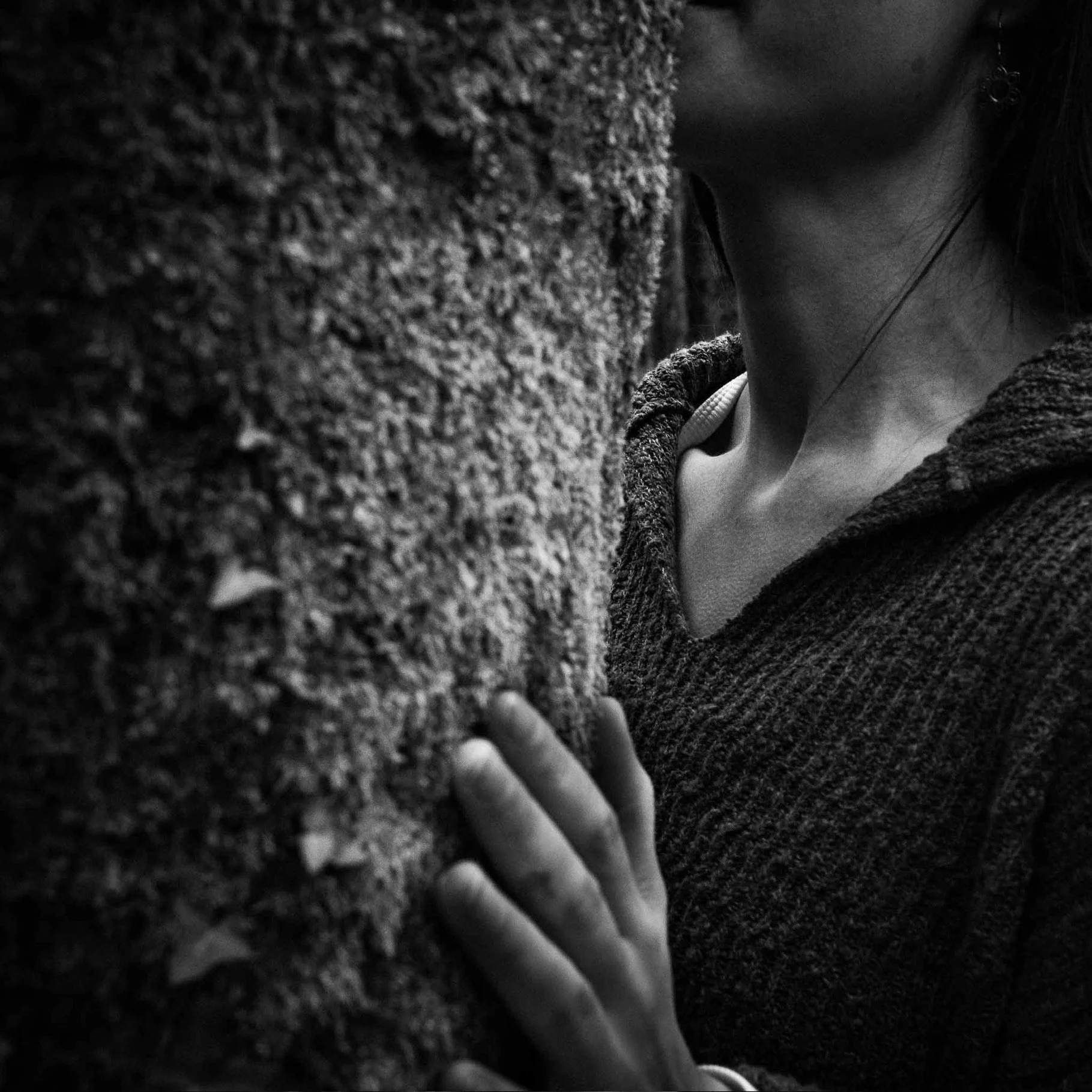 Close-up black and white photo of a person's face, showing only the lower part of their nose, lips, neck, and part of their shoulder. They are wearing earrings and a textured sweater, with a tree in the background.