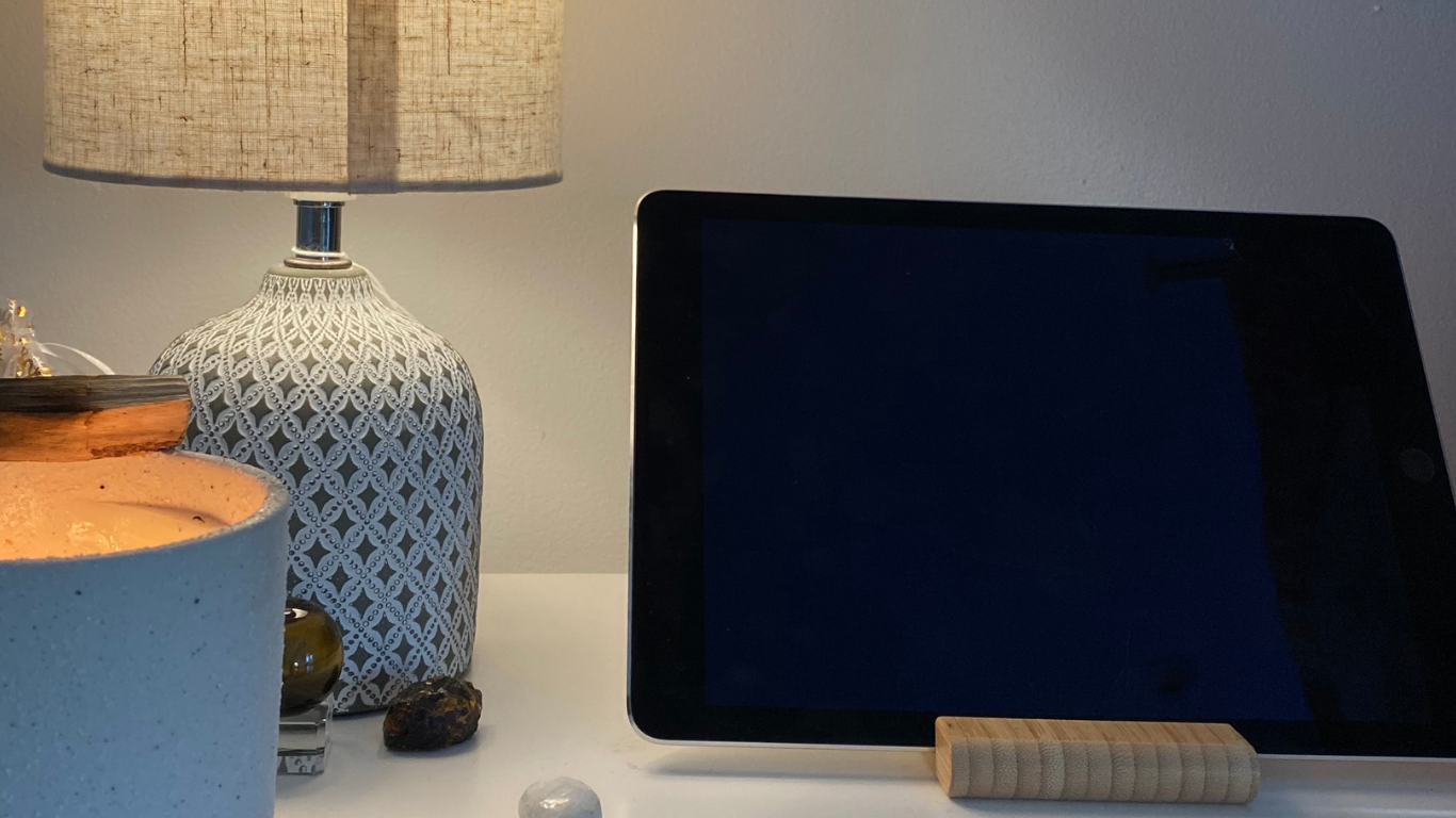 A white table with a large candle, a decorative vase with a lamp, a small yellowish object, and an electronic device on a wooden stand.
