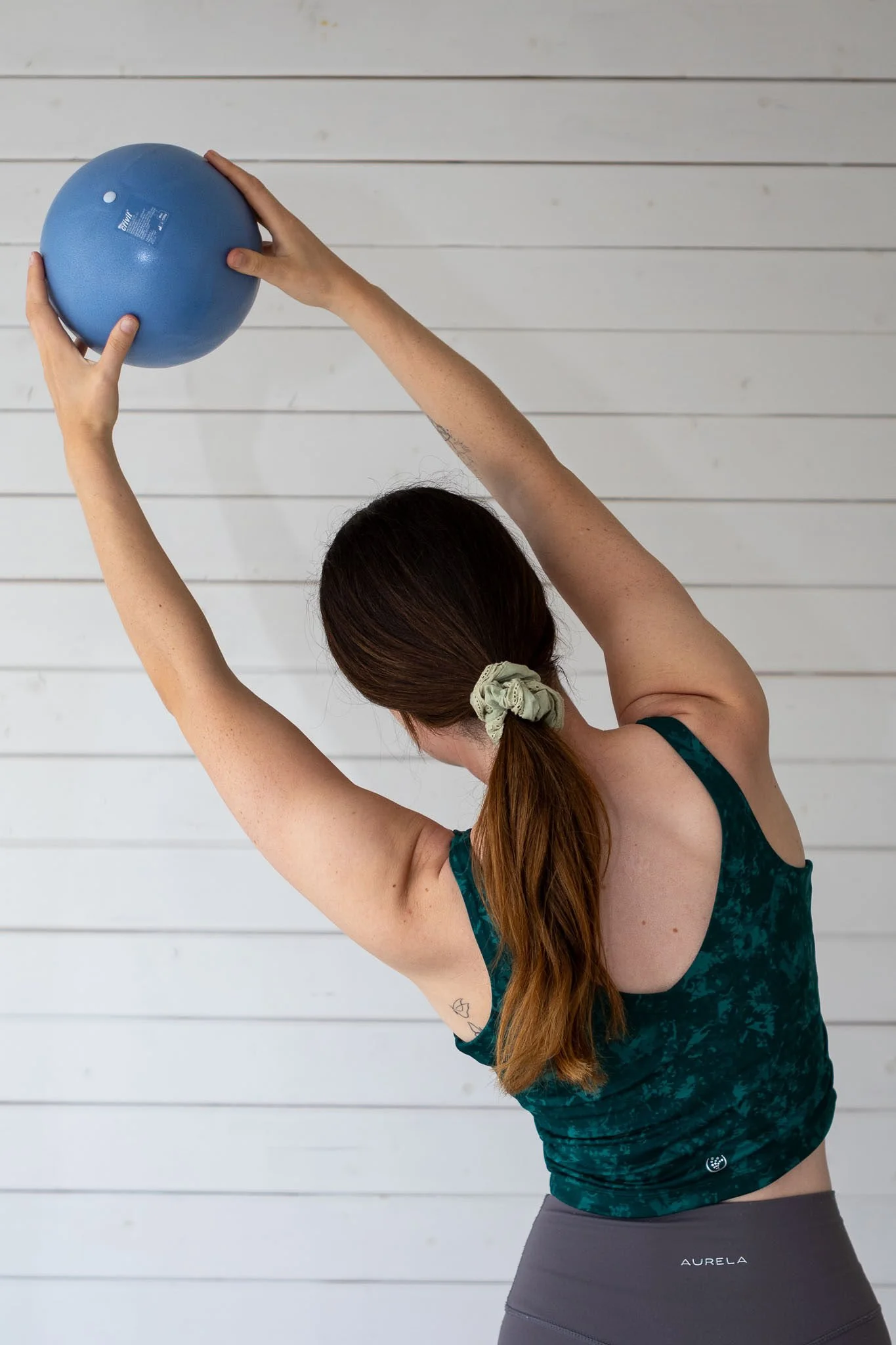A woman with long brown hair tied with a light-colored scrunchie is stretching her arms upward while holding a blue exercise ball. She is wearing a dark green patterned tank top and dark gray leggings. The background features a white wooden wall.