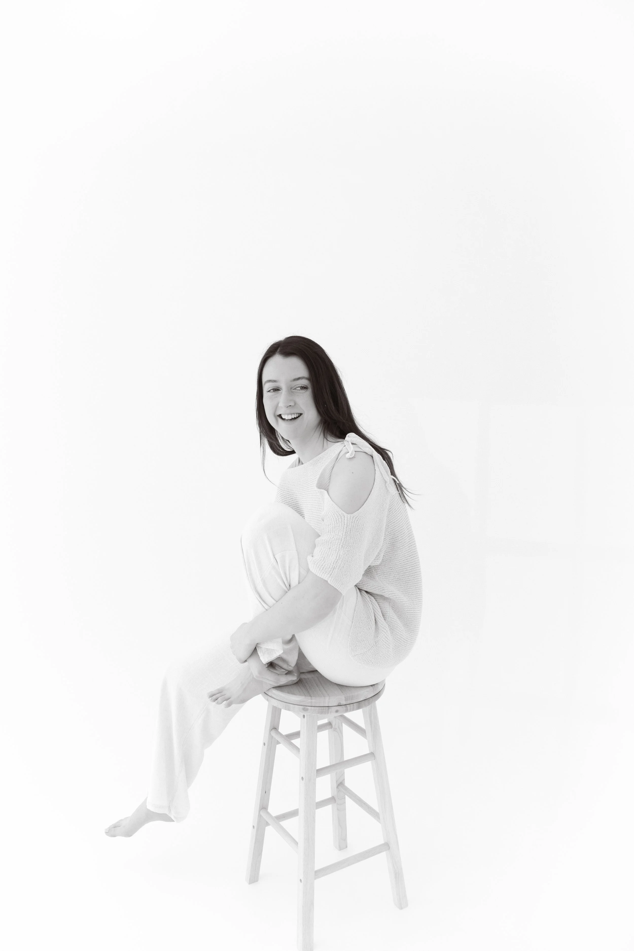 Black and white photo of a young woman sitting on a stool, smiling and looking slightly to the side, with her legs crossed and wearing a short sleeve knitted top with cutouts.