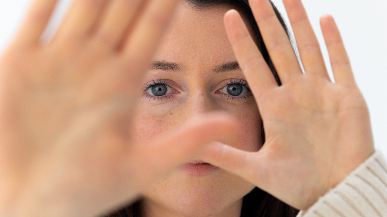 Close-up of a woman with blue eyes holding her hands up near her face as if peeking or shielding her eyes.