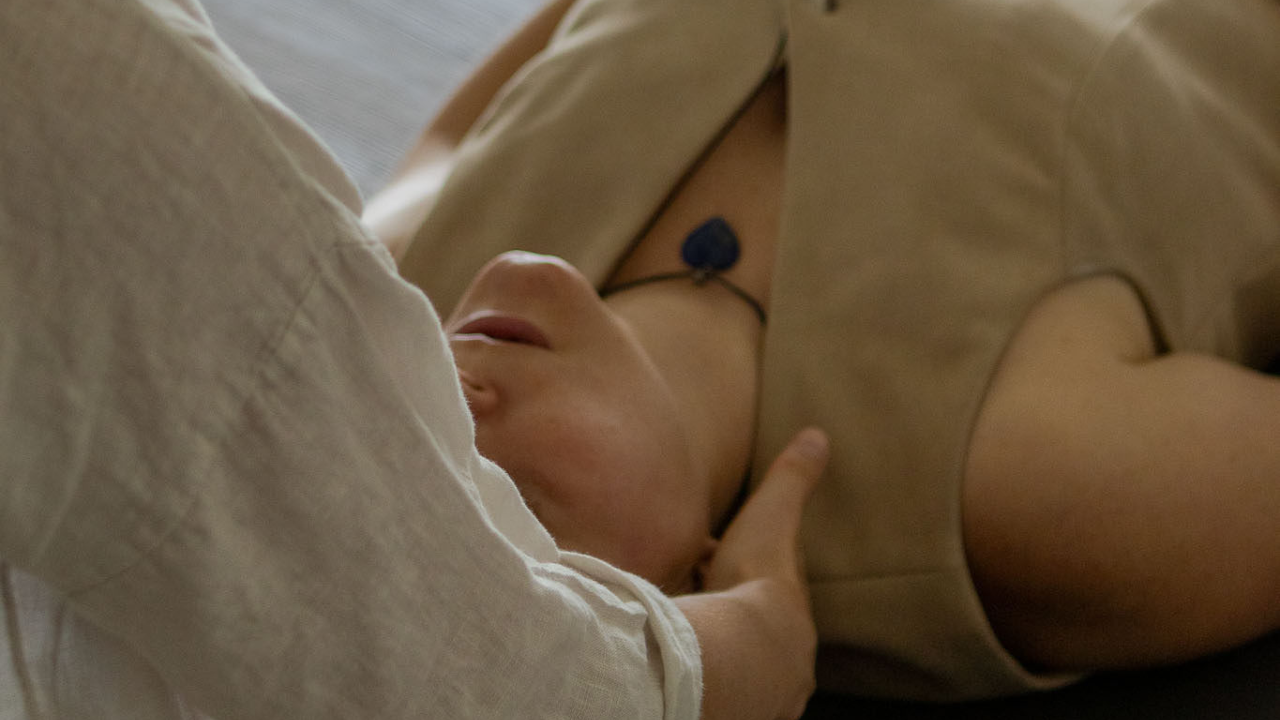 Person lying down with a hand on their face, wearing a beige shirt, visible chest with a necklace, in a relaxed or resting position.