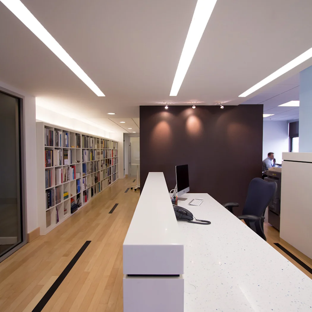 Modern office reception area with a white desk, black chair, computer, and a dark accent wall, with a bookshelf along the left side and a person working in the background.