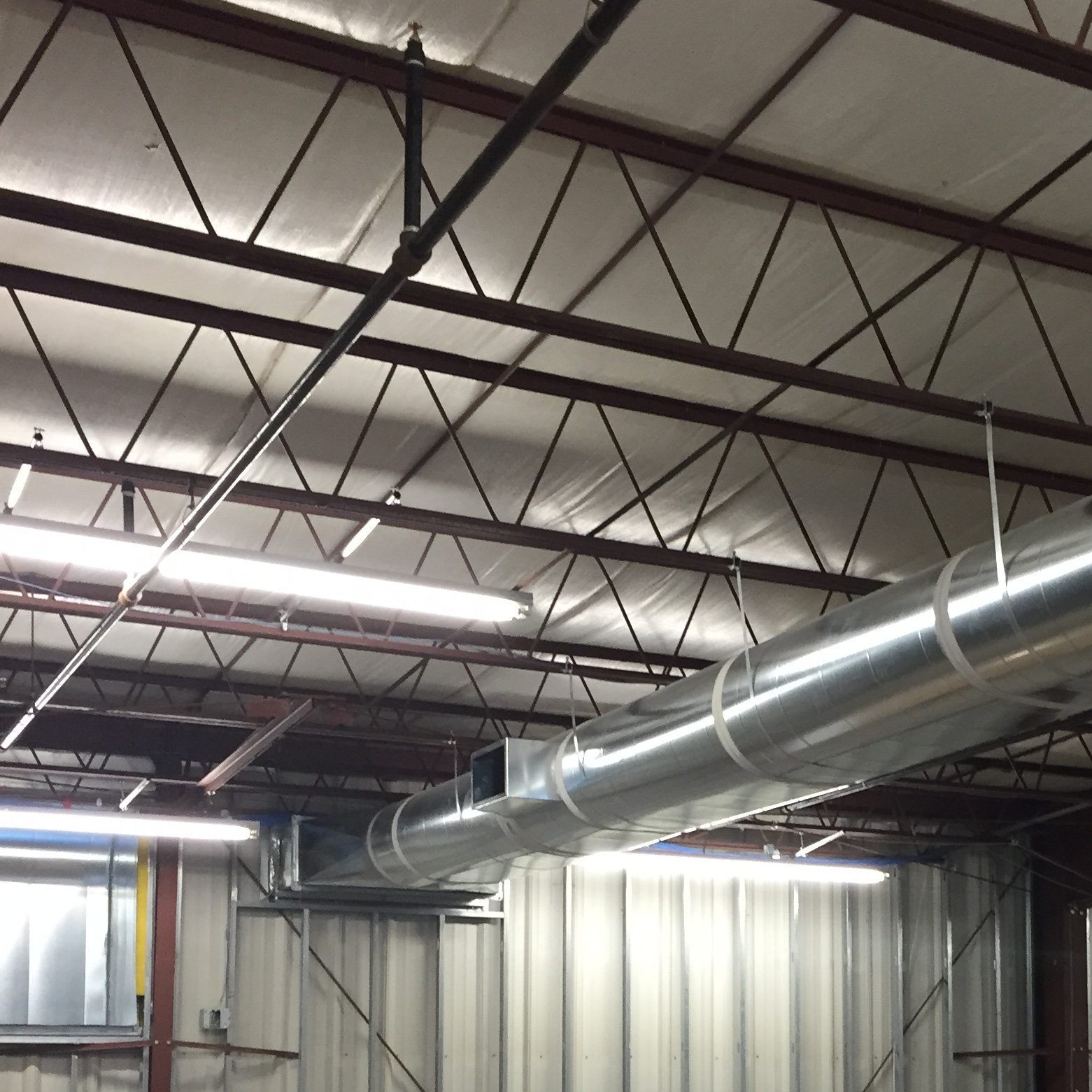 Interior view of an industrial or warehouse ceiling with metal beams, large silver air ducts, exposed pipes, and fluorescent lighting.