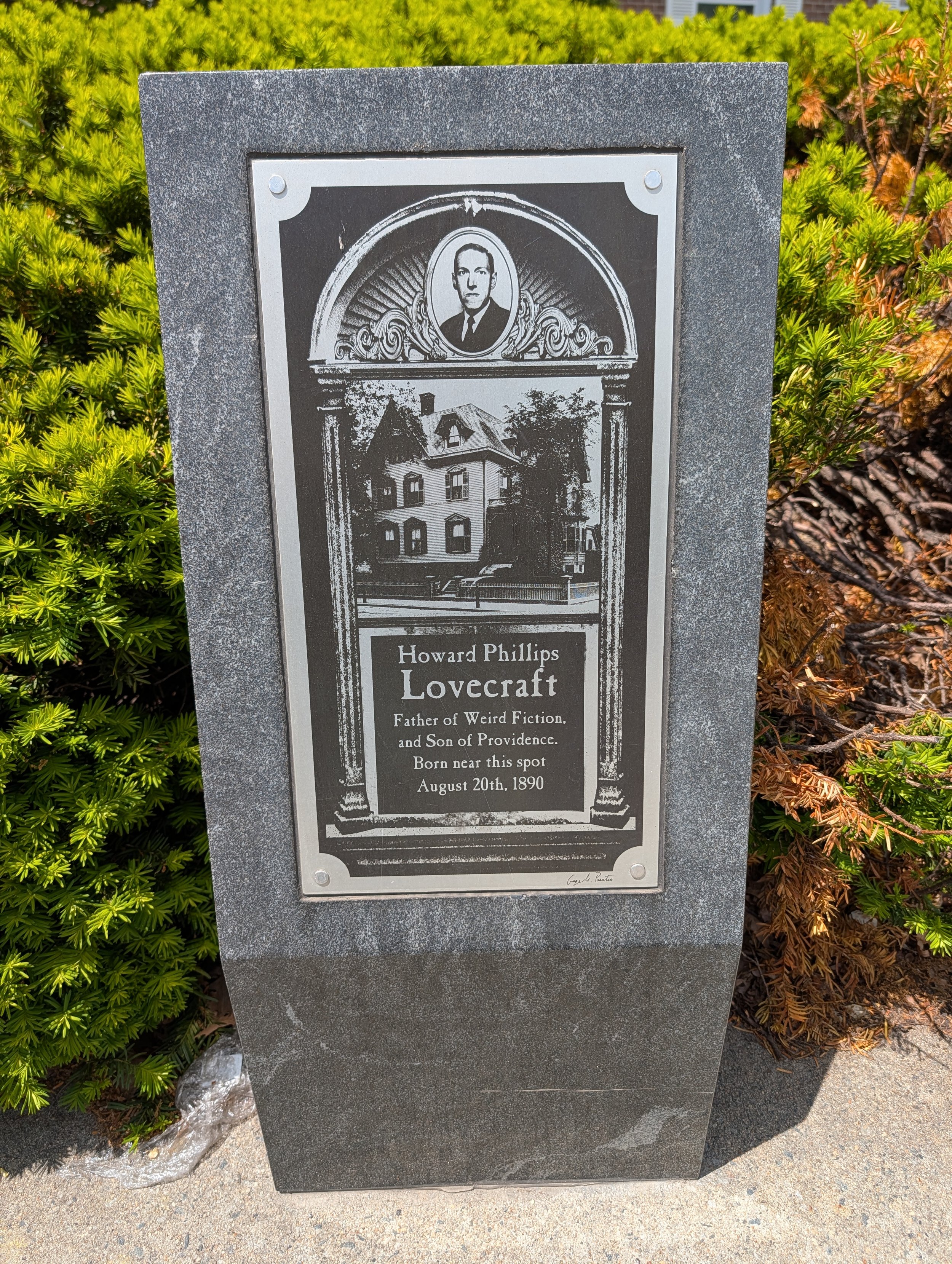 A memorial plaque dedicated to Howard Phillips Lovecraft, stating he was the father of weird fiction, born in Providence on August 20th, 1890, located outdoors among bushes and on a concrete sidewalk.