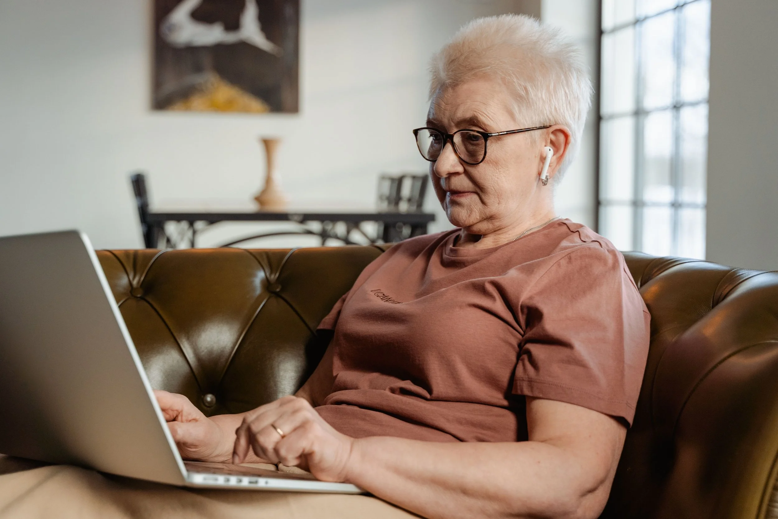 Ältere Frau mit weißen Haaren und Brille nutzt ein Laptop und trägt kabellose Kopfhörer im Wohnzimmer.