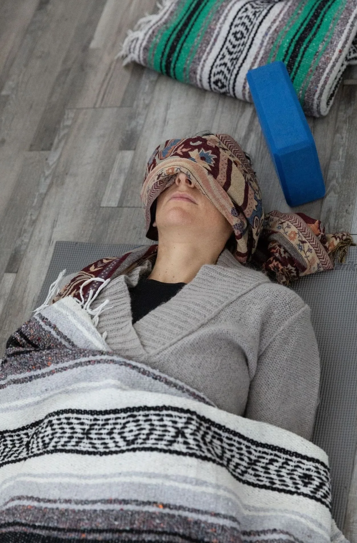 Person lying on the floor with a cloth covering their eyes, wearing a patterned sweater during savasana.