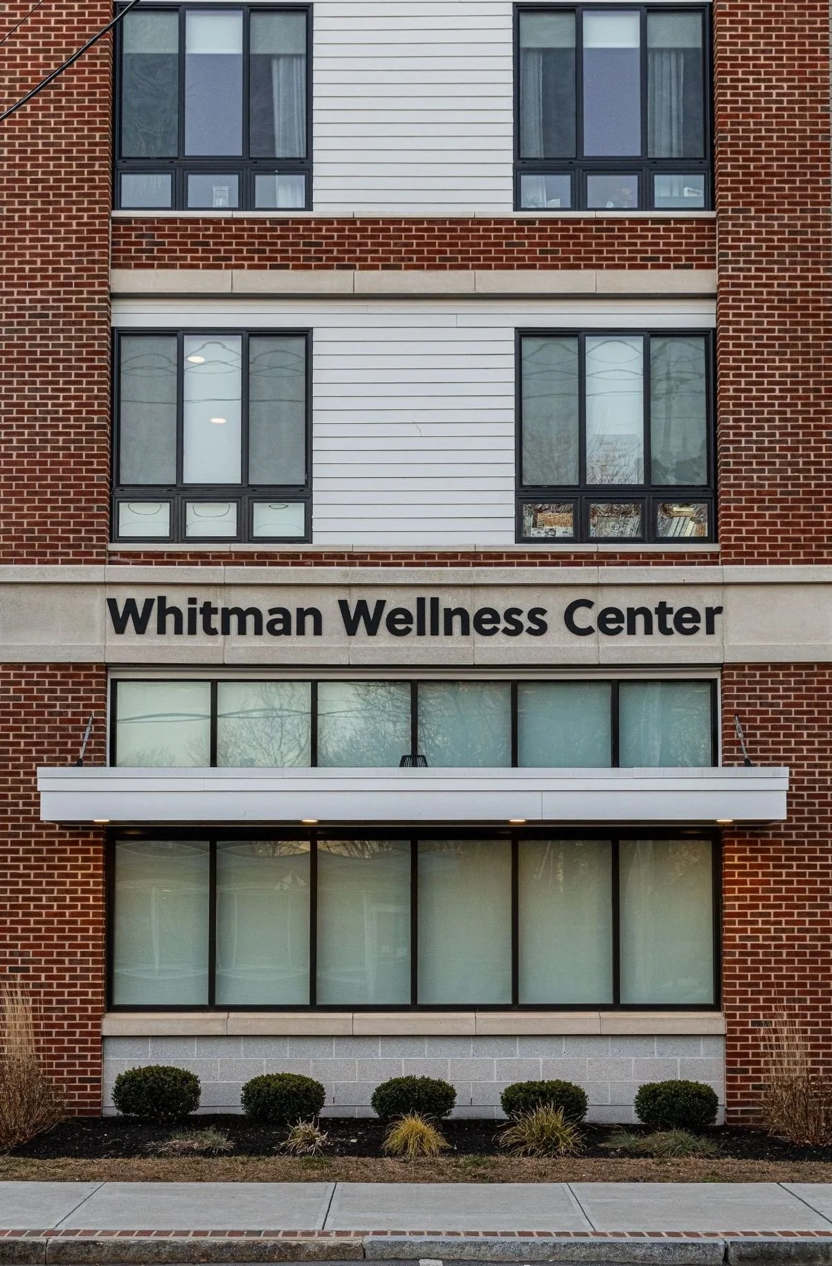 Front view of a multi-story building with a sign that reads 'Whitman Wellness Center'. The building has a combination of red brick, white panels, and large glass windows. The lower level has a row of shrubs and a sidewalk.