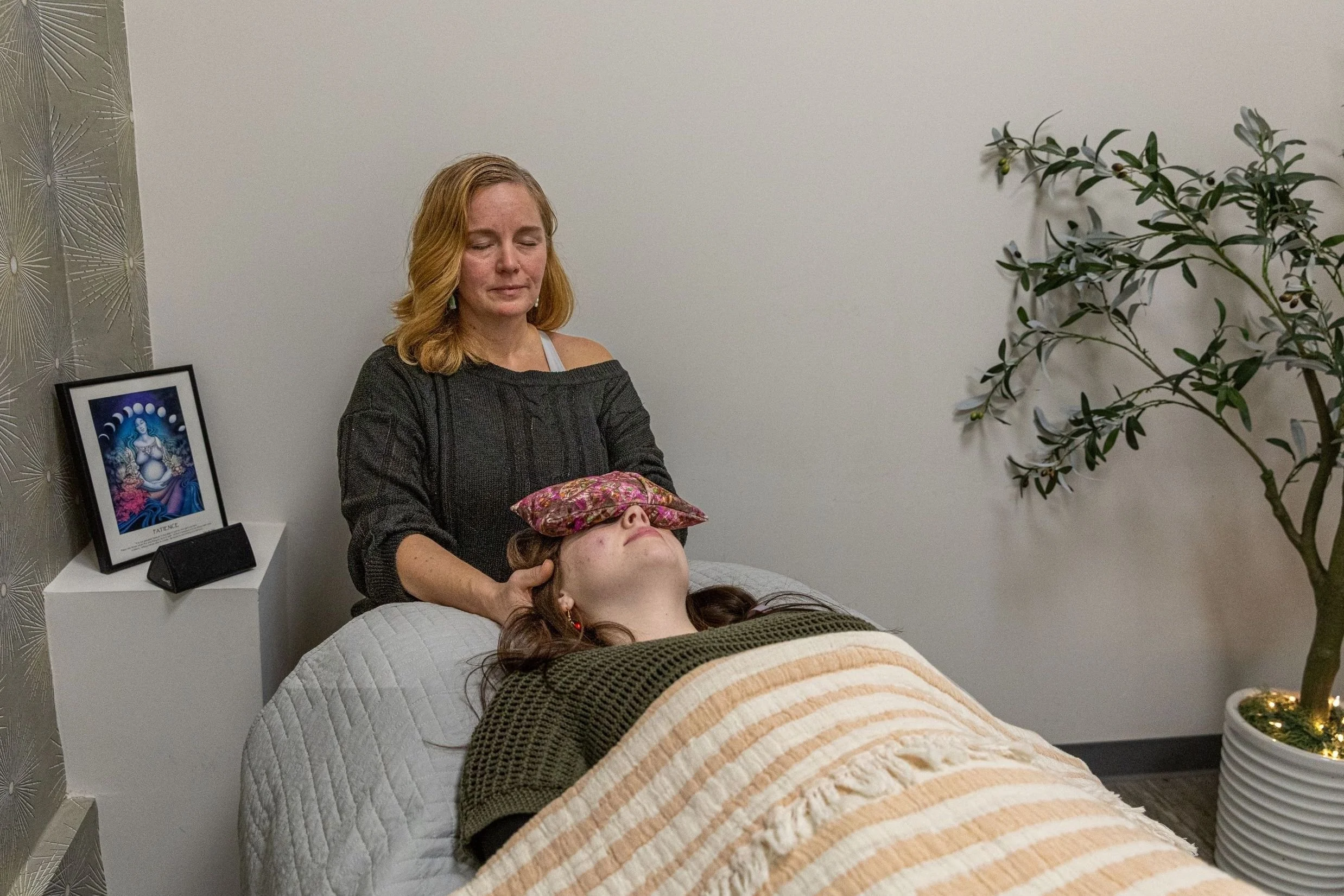 A woman lying on a bed with her eyes closed, receiving reiki from another woman in a room decorated with a framed artwork, a side table, and a potted plant.