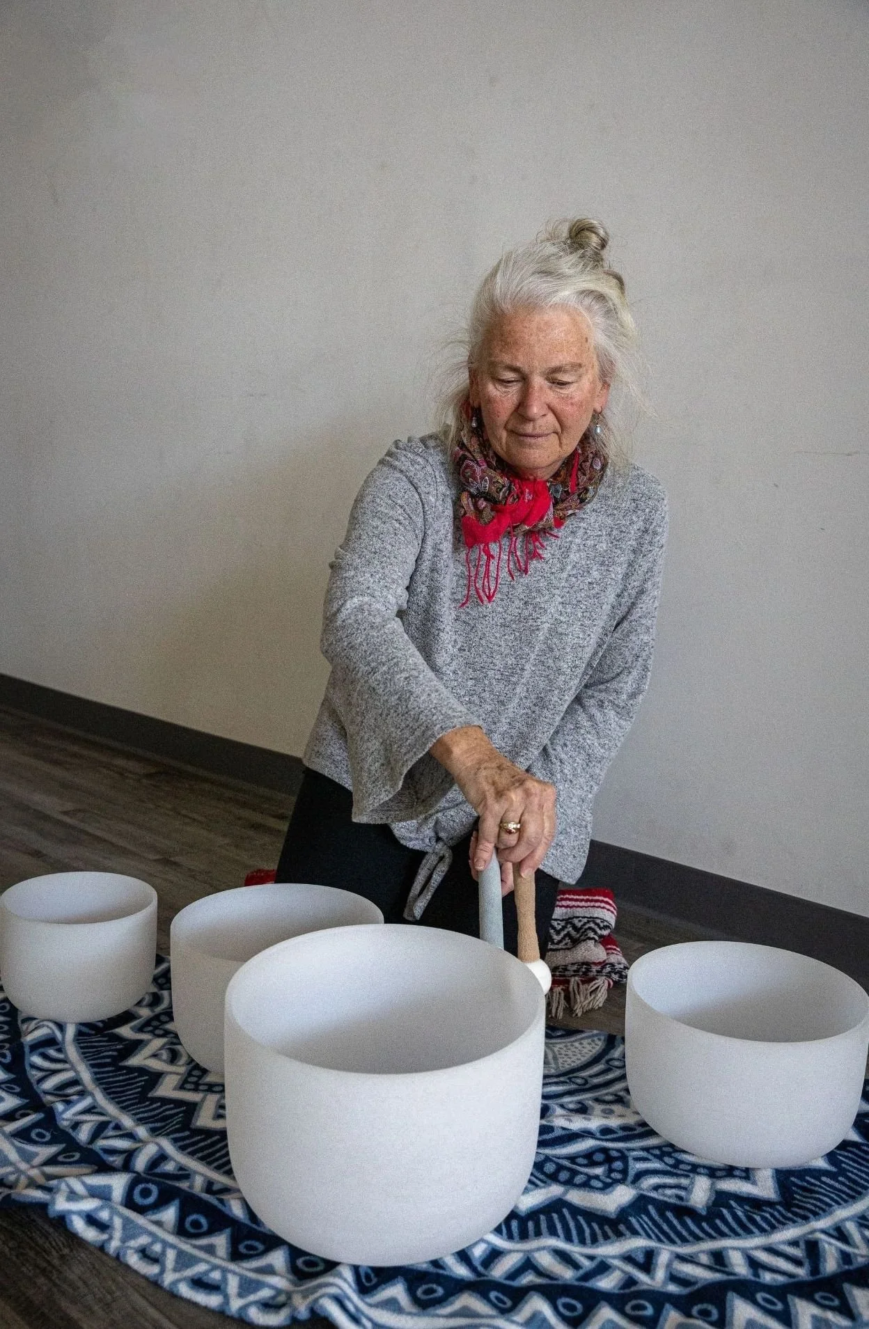 Older woman with gray hair and a colorful scarf playing crystal singing bowls on a patterned cloth.