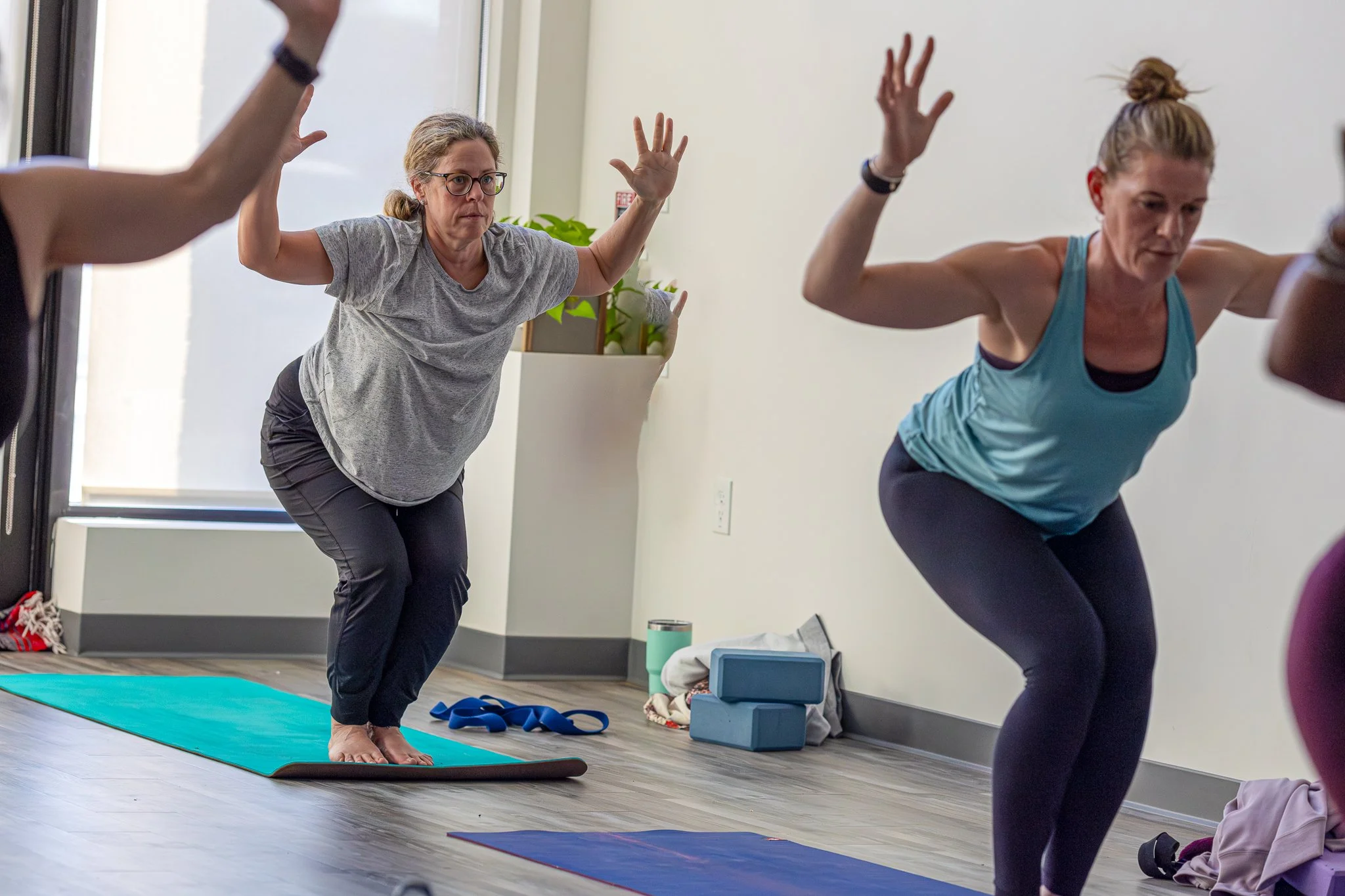 Two women are practicing yoga or a similar exercise in a room with wooden floors and large windows. One woman in a gray T-shirt and glasses is balancing on a yoga mat, with her arms raised. The other woman in a blue athletic top is kneeling and also 
