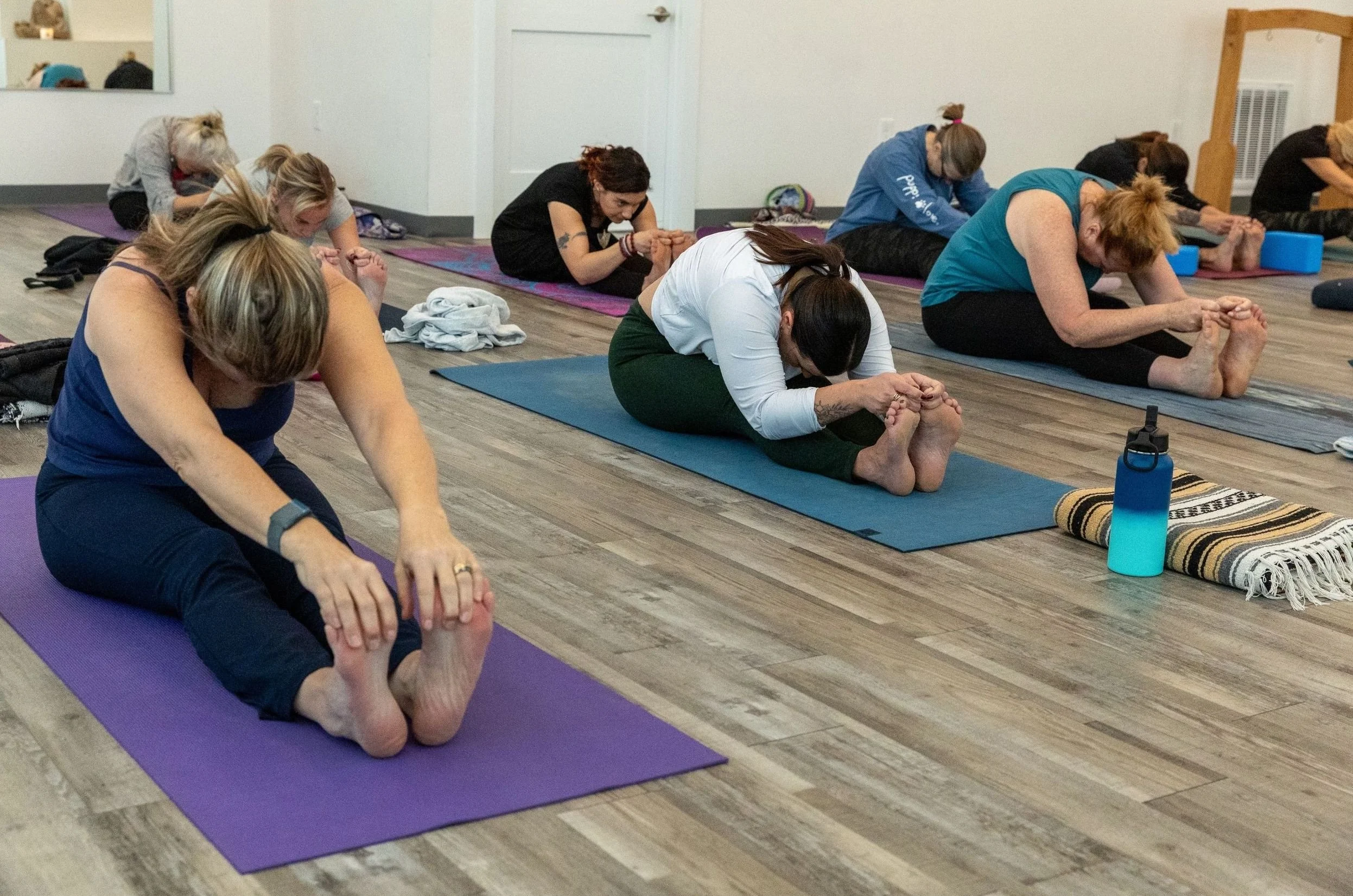 A group of women participating in a yoga class, sitting on mats and bending forward to hold their feet, with their heads down and hands on their feet.