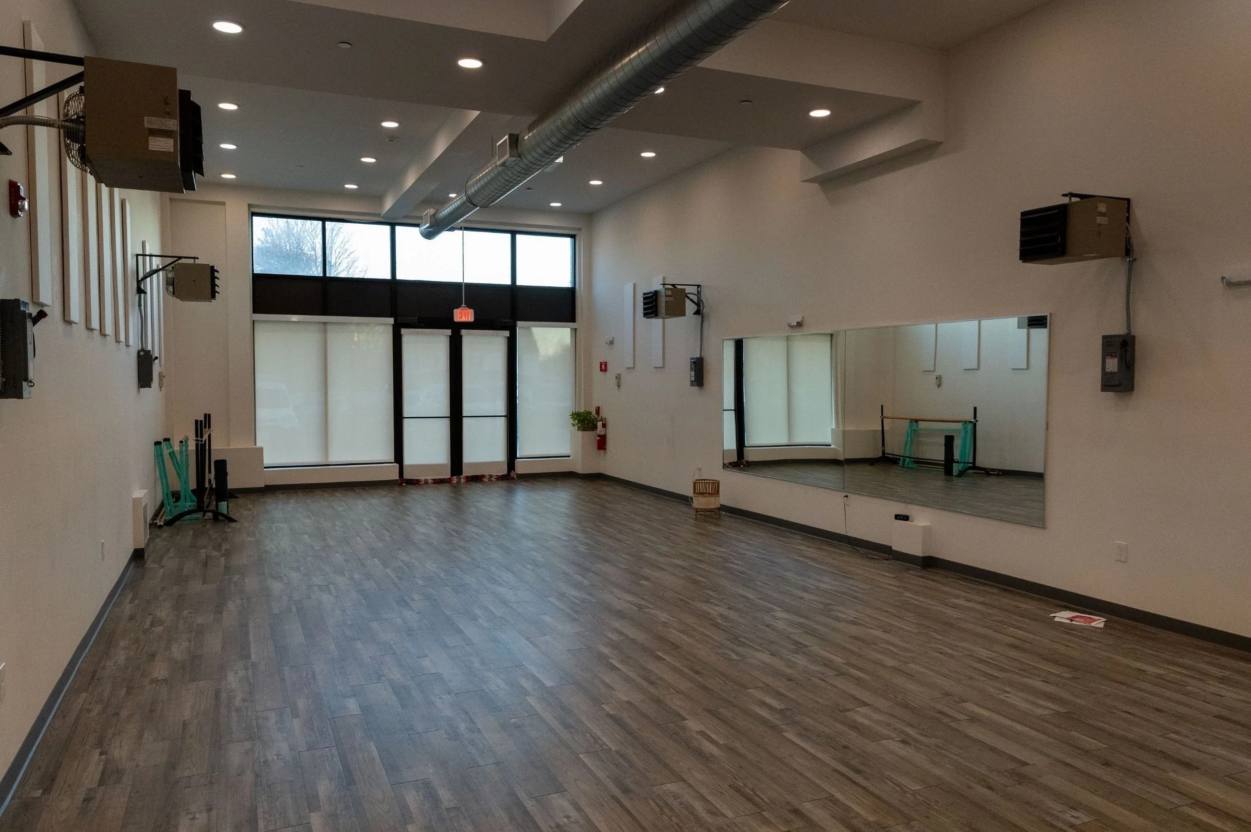 Empty commercial or fitness studio with wooden flooring, large glass windows, ceiling lights, wall-mounted speakers, and a large mirror.