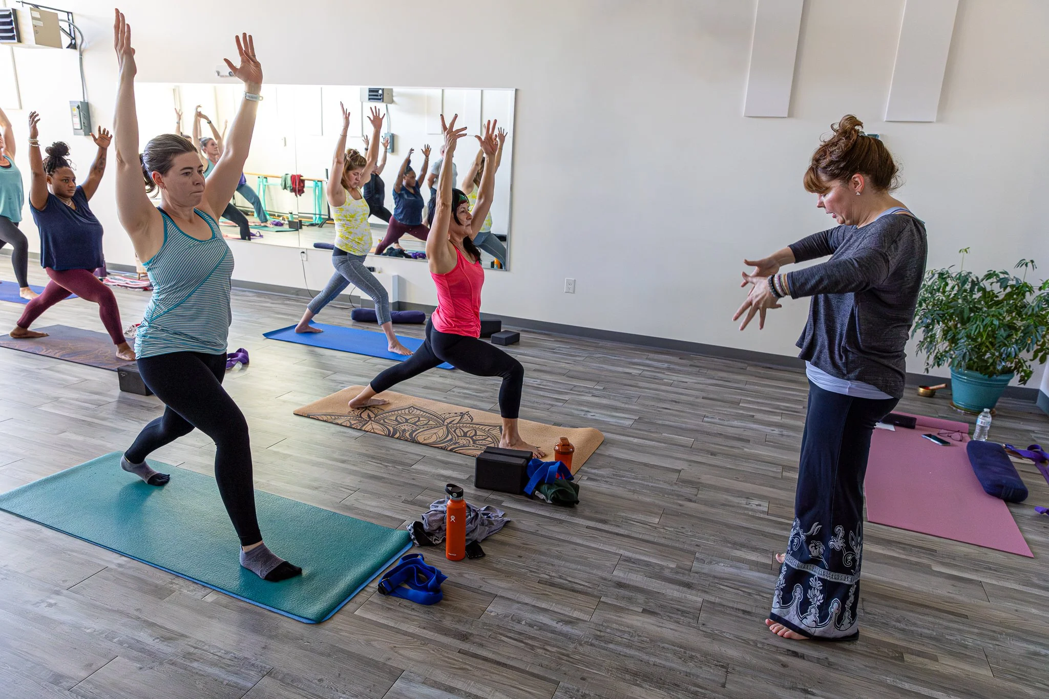 A group of women practicing yoga in a studio, following an instructor who is leading a pose. The women are on yoga mats, raising their arms, with a mirror on the wall reflecting their movements. The studio has light-colored walls, wooden flooring, an
