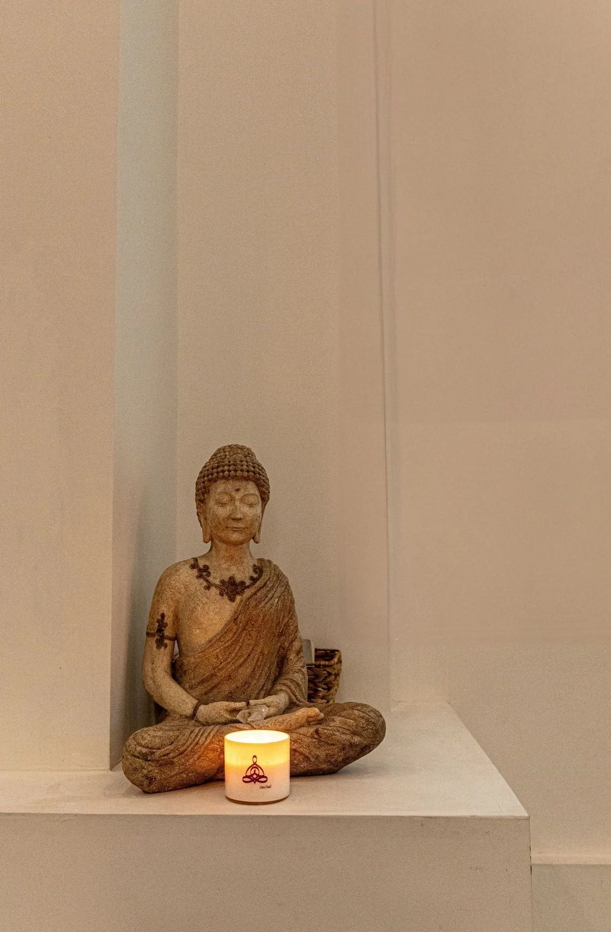Buddha statue with a calm expression, seated with crossed legs, wearing a necklace and draped in a robe, with a lit candle in front of it on a white surface, in a minimalistic room.