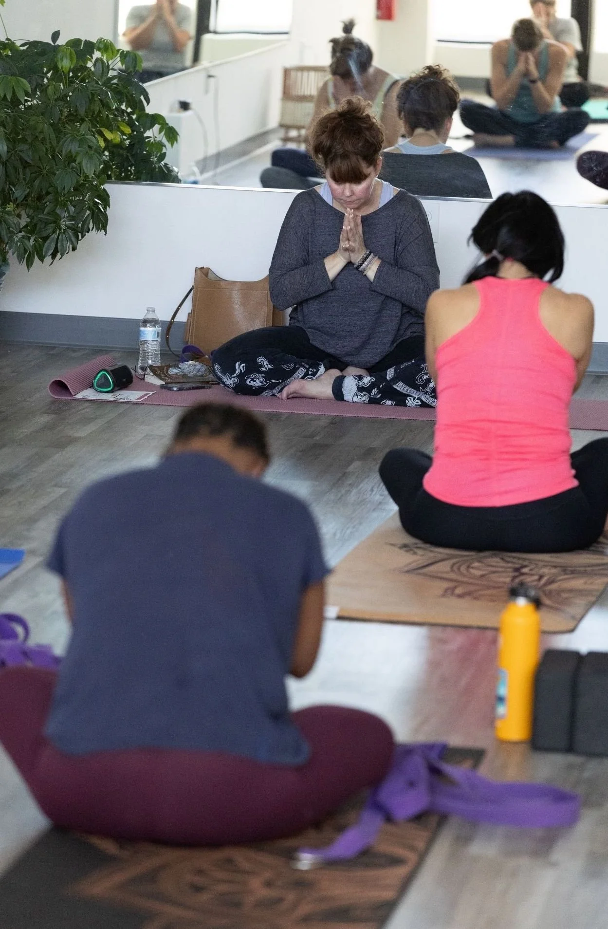 People practicing yoga or meditation in a studio, sitting cross-legged on mats with hands in prayer position, some with eyes closed and others looking down.
