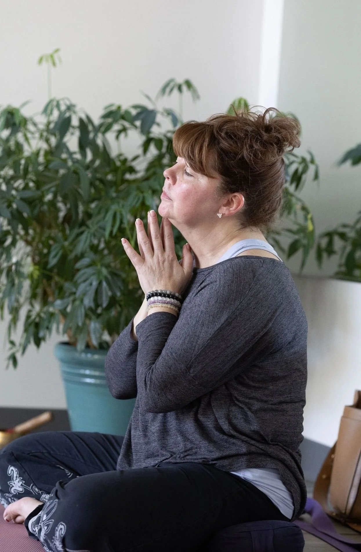 A woman practicing yoga or meditation, sitting cross-legged in a peaceful pose with eyes closed and hands in prayer position near her face, indoors with a large potted plant in the background.