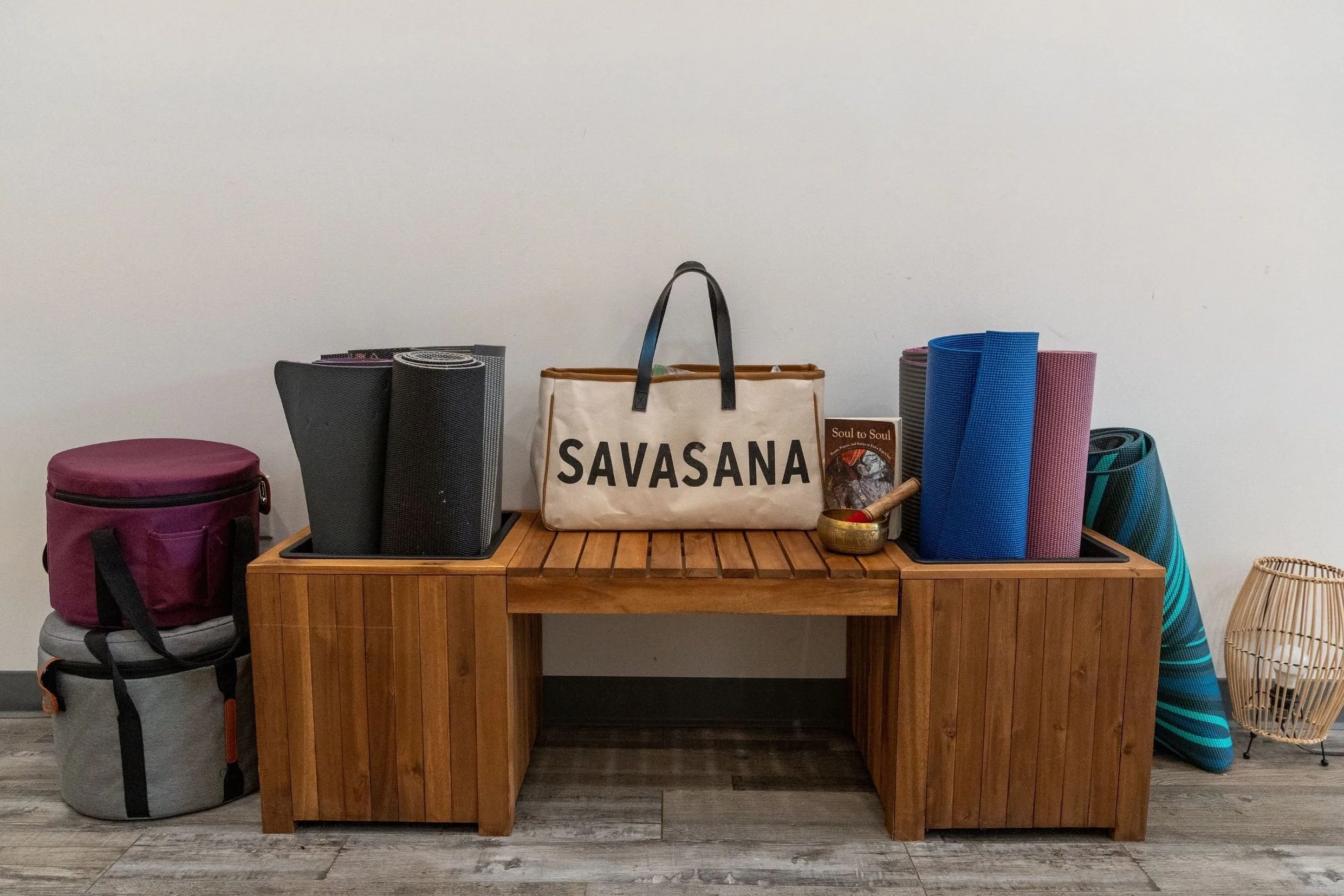 A wooden table holding rolled yoga mats, a bag, and other items. Behind, a pink bag and a gray backpack are on the floor. To the right, a small rattan lantern and a framed picture are visible. The background is a plain white wall.