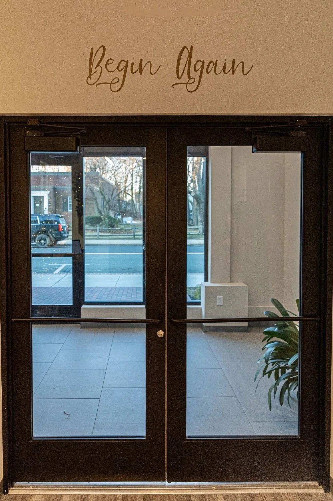 Double glass doors with a sign above that says 'Begin Again'. Through the doors, a sidewalk and street with parked cars and trees are visible, along with some indoor plants.