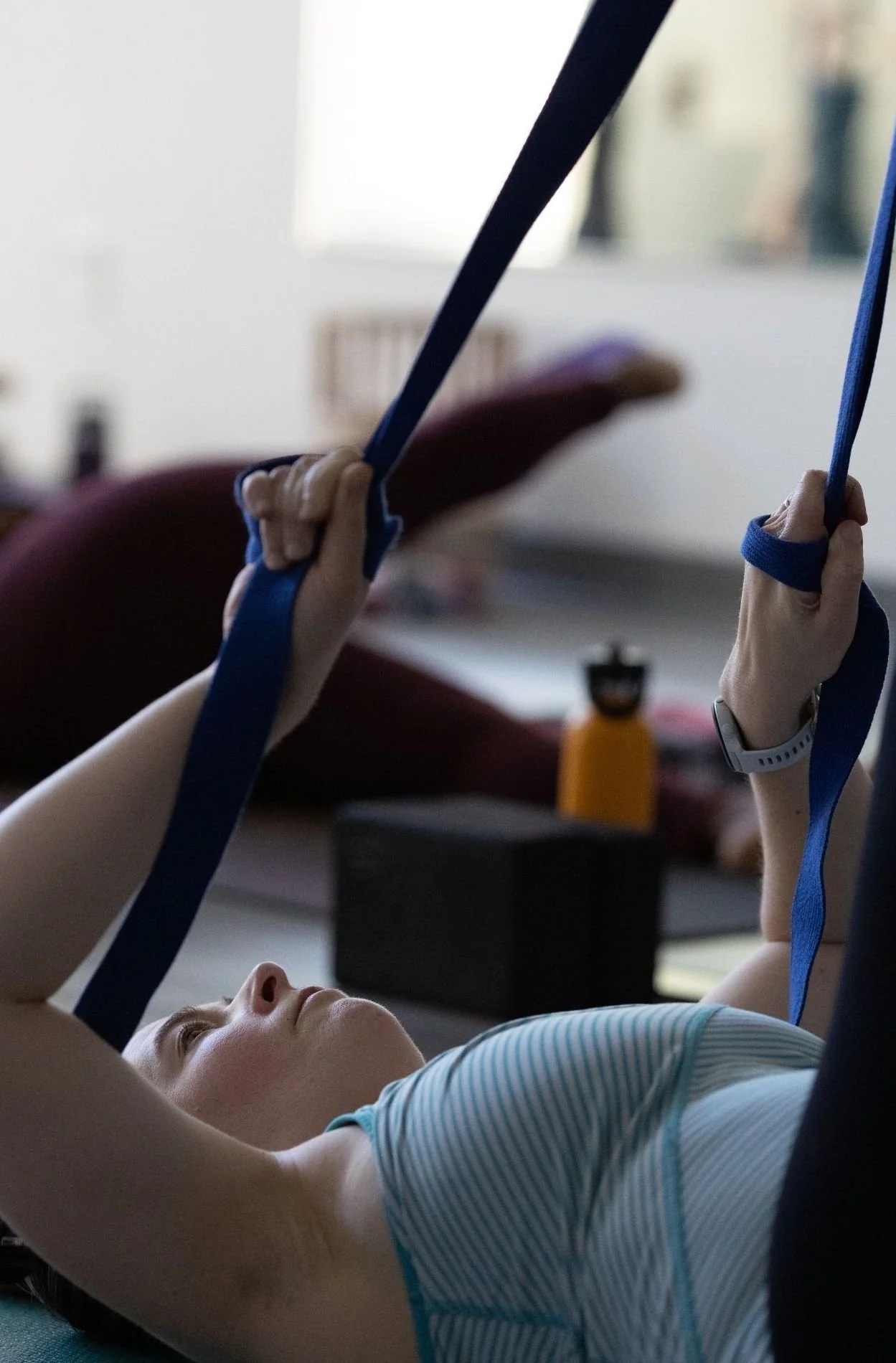 A young girl lying on her back on a workout mat, lifting her arms, holding resistance bands, and exercising indoors.