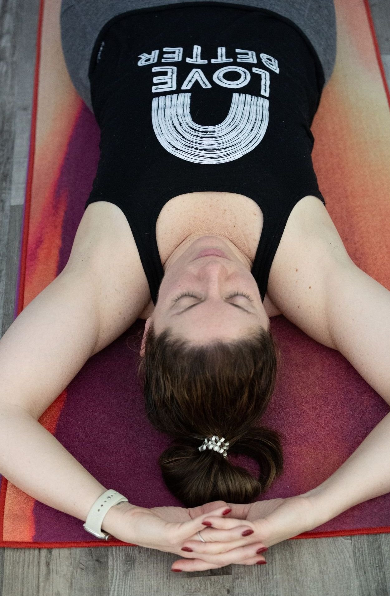 A woman lying on her back on a yoga mat with her eyes closed and arms resting above her head, wearing a black tank top with white lettering and a graphic design, and a white watch on her left wrist.