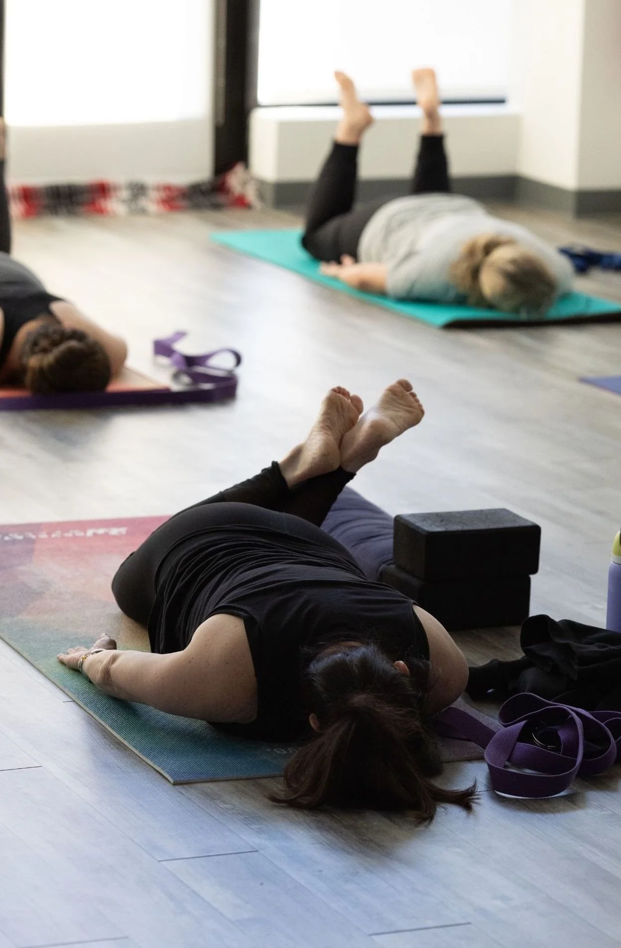 A group of people practicing yoga in a studio, lying on colorful yoga mats, with one person in the foreground, and another person in the background raising their legs while lying on their stomach.