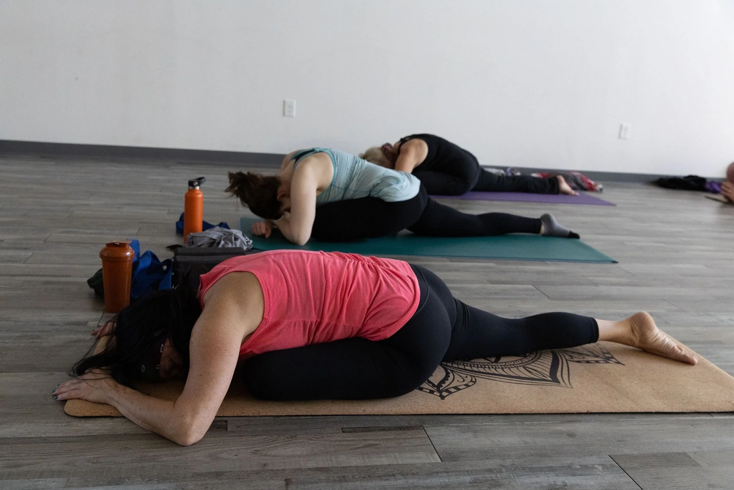 Three women are practicing yoga on mats in a studio with a plain white wall and wooden floor. They are in a forward bend pose with their heads resting on their legs, and each woman has beverages and personal items nearby.