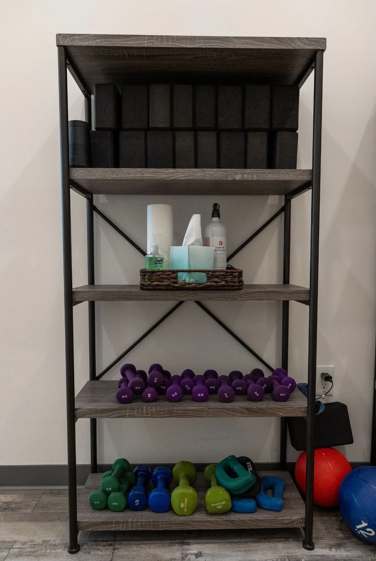 A black metal and wood shelving unit with four shelves holding workout equipment, including dumbbells, kettlebells, a medicine ball, foam rollers, and a basket with toiletries.
