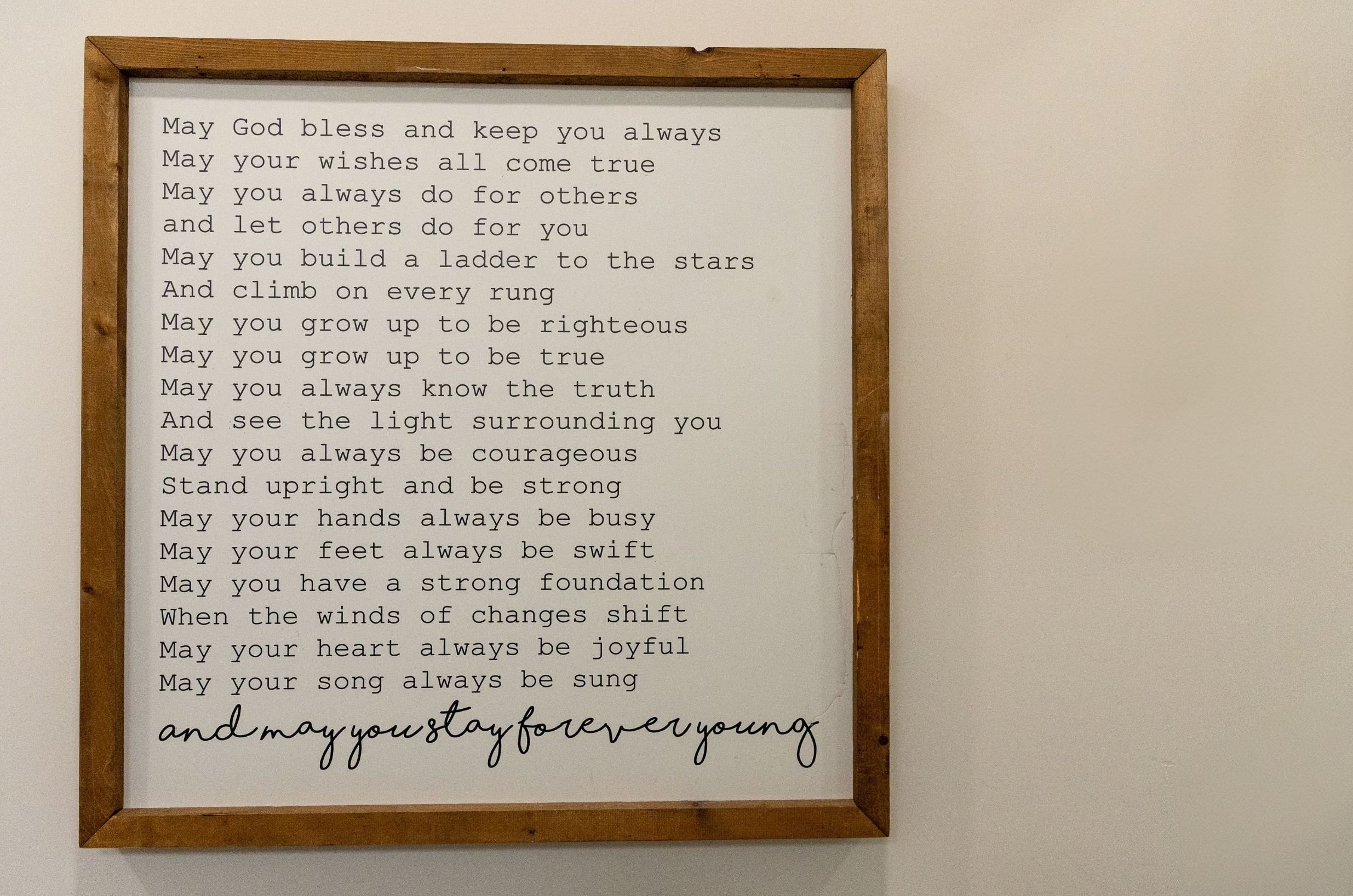 A framed printed poem with a wooden border on a plain wall. The poem offers blessings and encouragements, including prayers for strength, courage, and joy, ending with "and may you stay forever young."