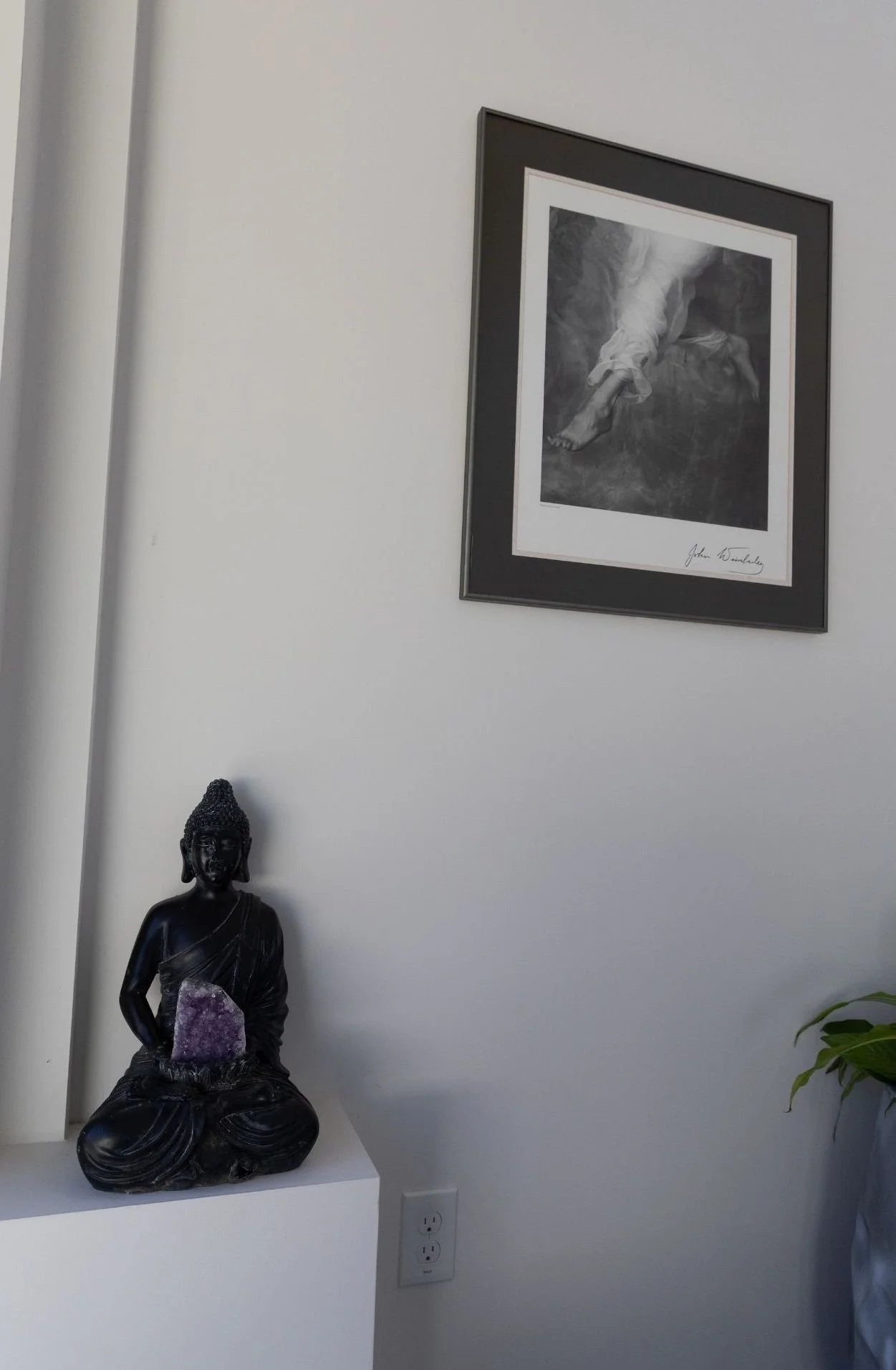 A black Buddha statue holding a purple amethyst crystal, placed on a white stand against a white wall, with a framed black-and-white religious artwork and a potted plant partially visible.