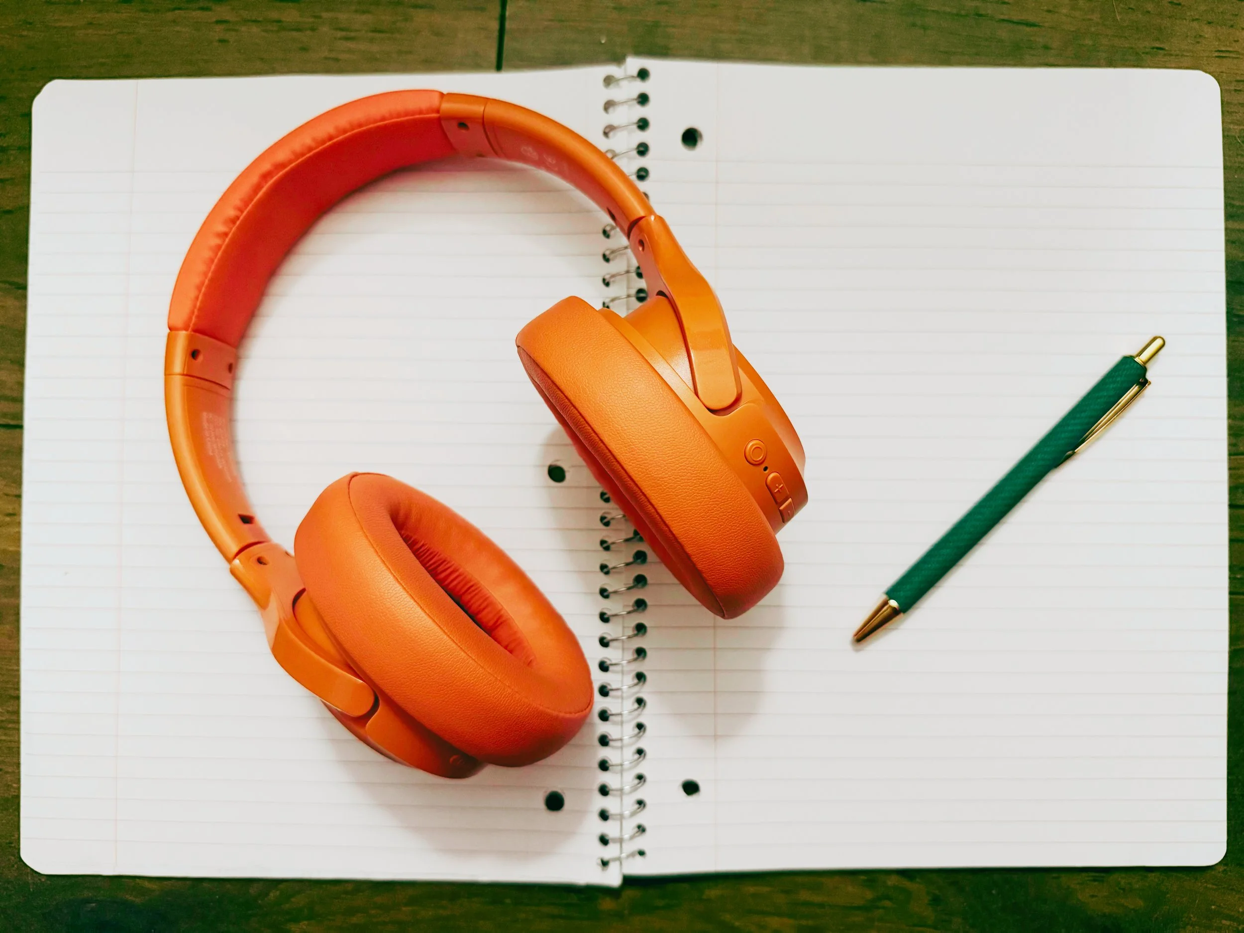 Orange over-ear headphones, a green pen with gold tip, and a spiral notebook with blank ruled pages on a wooden surface.