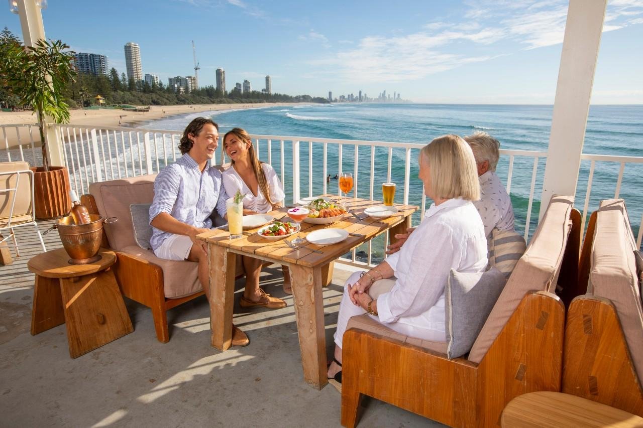 Four people enjoying a meal together on a balcony with ocean view, a city skyline in the distance, and clear skies.