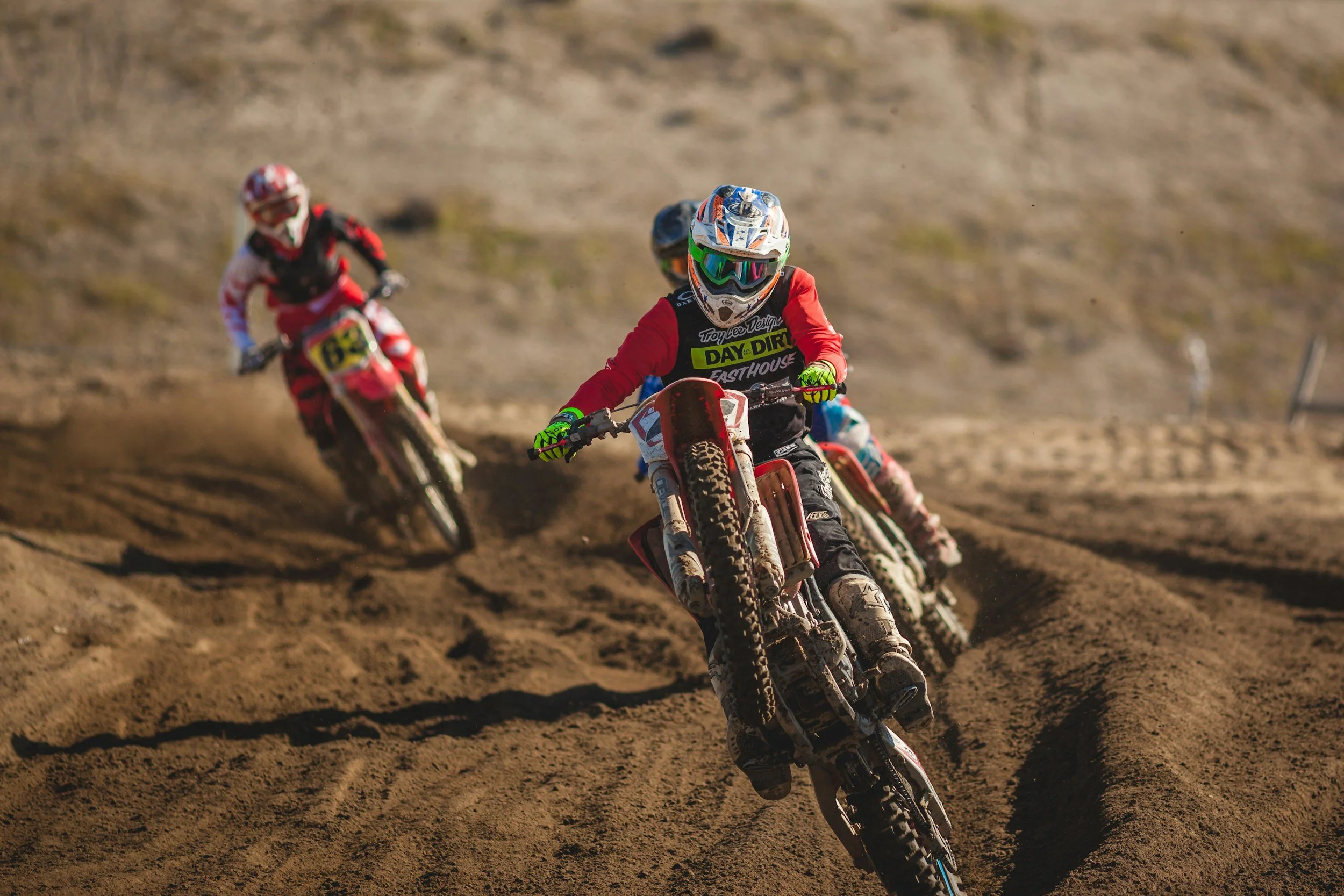 Three motocross riders racing on a dirt track with dust clouds, wearing colorful gear and helmets, leaning into a turn