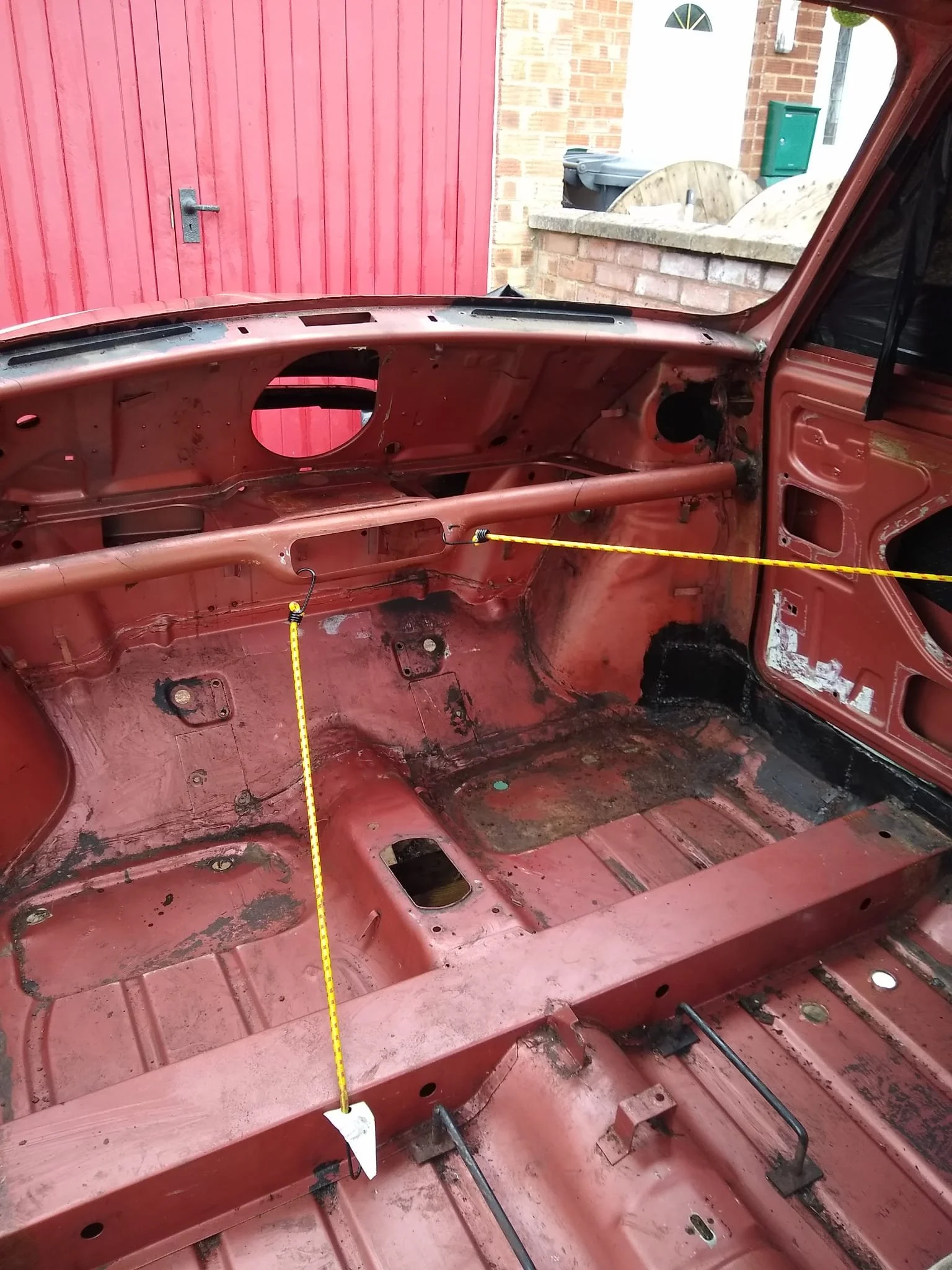 Interior of a vintage red car stripped of seats and interior panels, showing the bare metal floor and dashboard.
