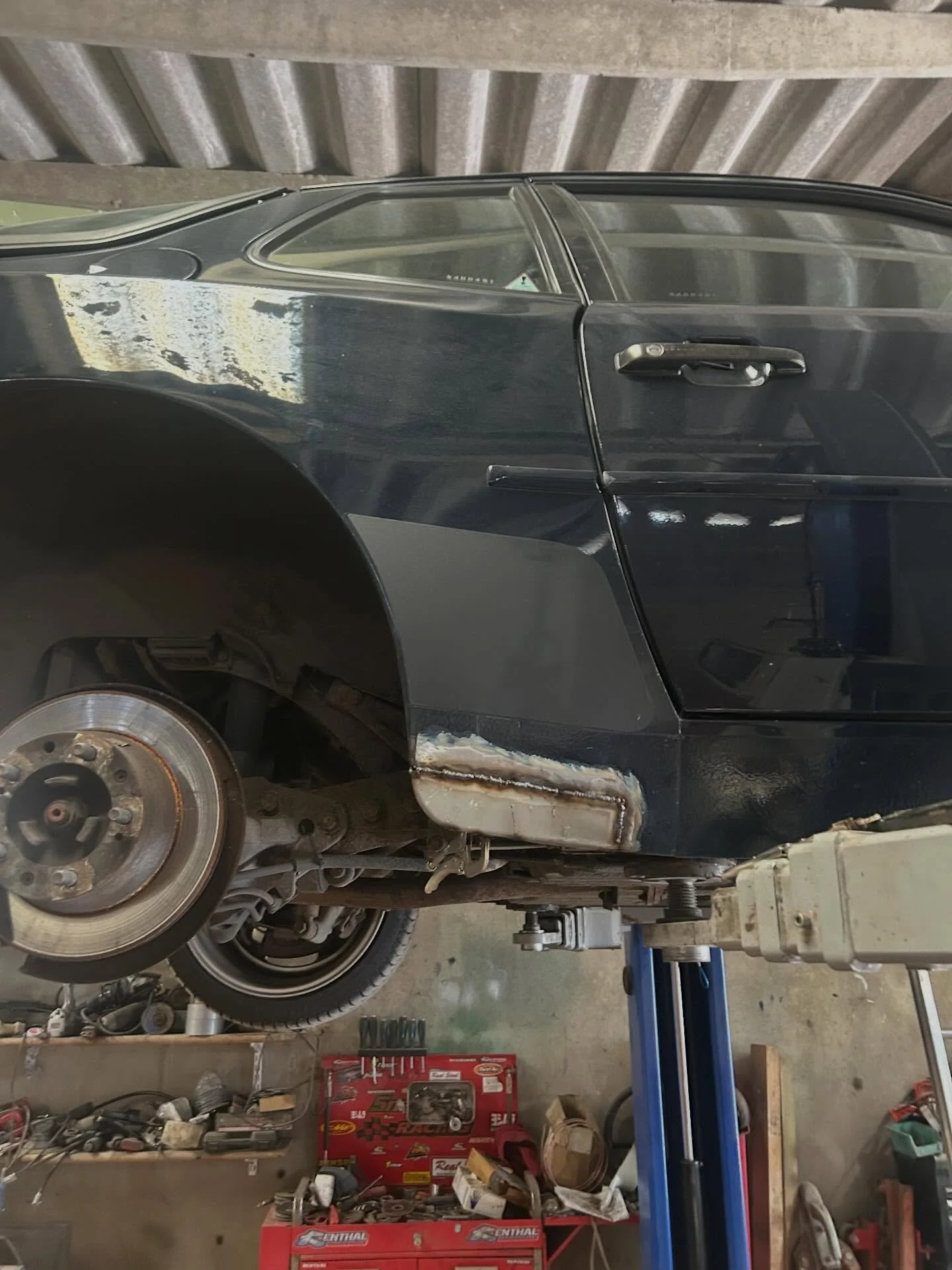 Black car on a hydraulic lift in an auto repair shop, with the front left wheel removed, exposing the brake rotor and suspension components. The workshop has various tools and equipment, including a red tool chest and shelves with parts and supplies.