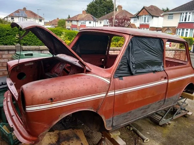 A rusty, orange station wagon with an open hood and black plastic covering the windows, parked outdoors on wooden blocks with houses in the background.