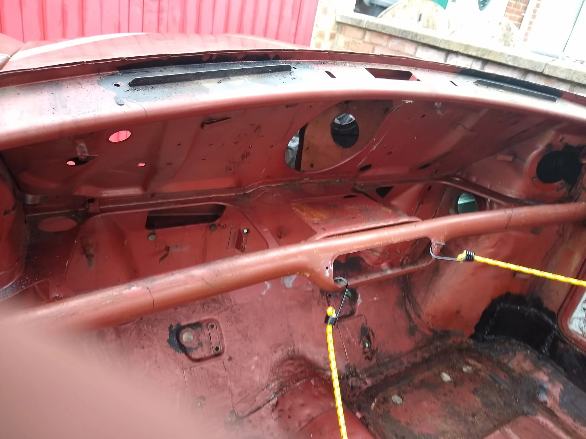 The interior of a vintage car painted in red, showing the dashboard and floor area with visible metal framework and wiring, possibly undergoing restoration.