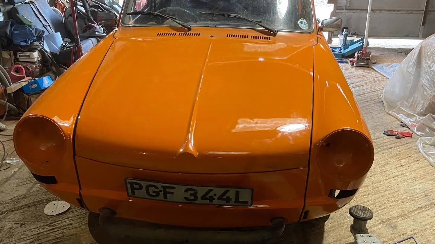 Front view of a vintage orange car in a garage, partially disassembled with a license plate PGF 344L, surrounded by tools and car parts.