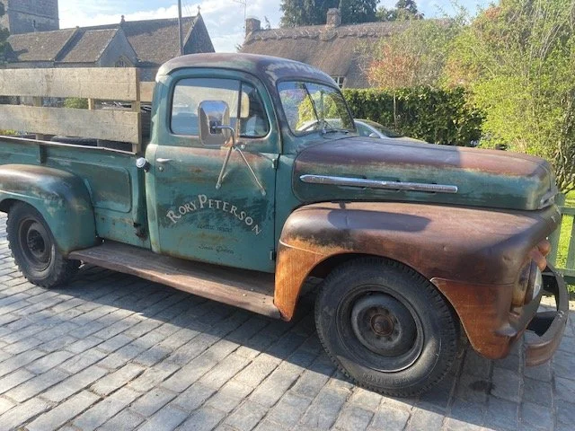 An old, rusty pickup truck with faded blue and brown paint, parked on a cobblestone driveway with a wooden fence and houses in the background.