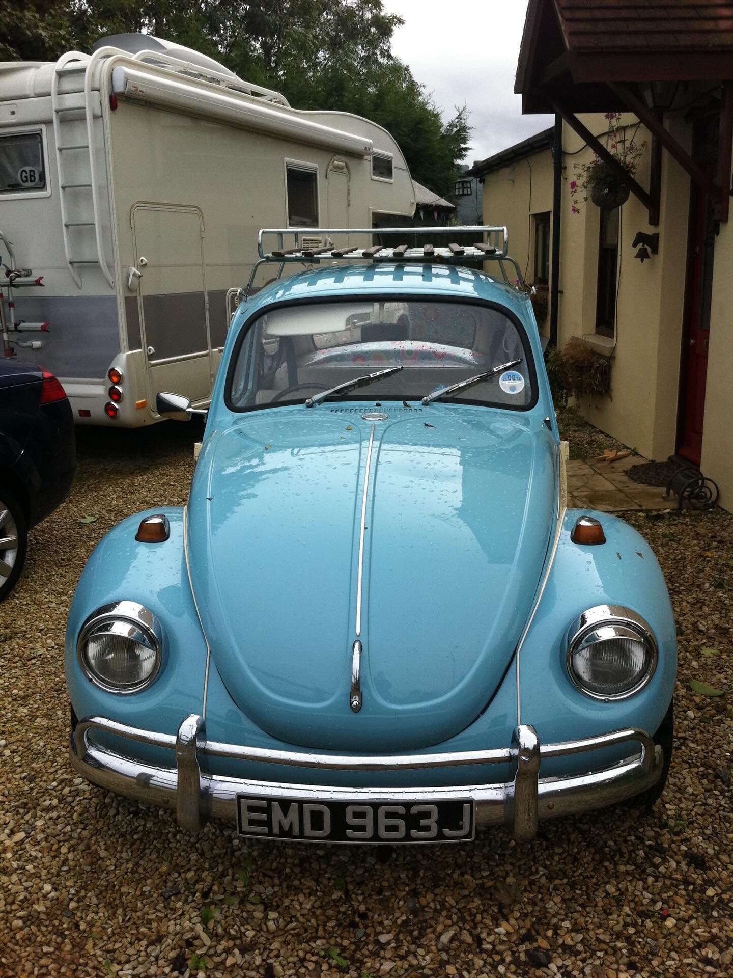 A vintage light blue Citroën 2CV car parked on a gravel driveway, with a white caravan and a house in the background.