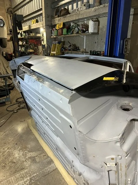 A partially assembled car body in a garage or workshop with tools on shelves in the background.