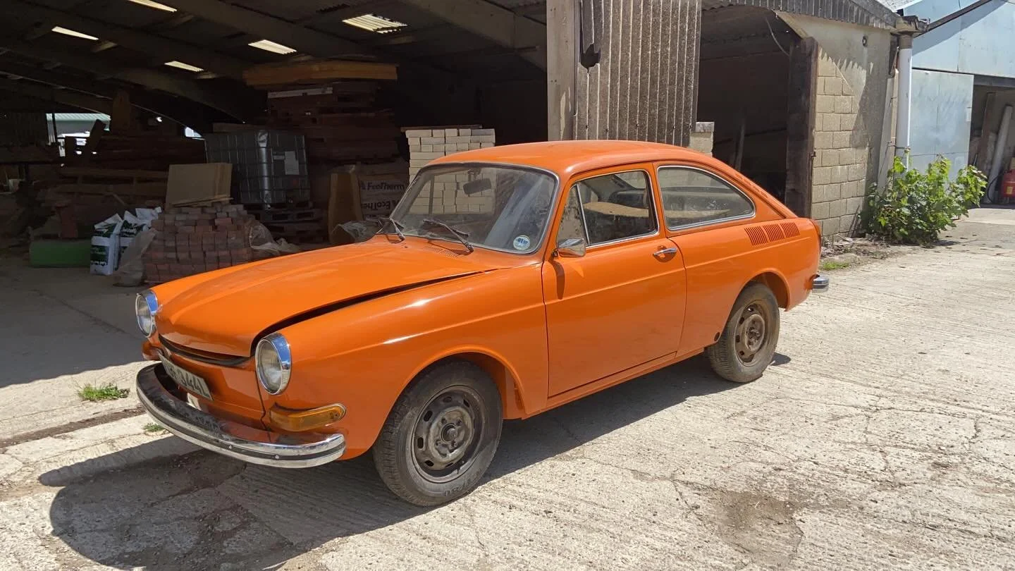 An orange vintage car parked outside a workshop or garage with building materials and plants in the background.