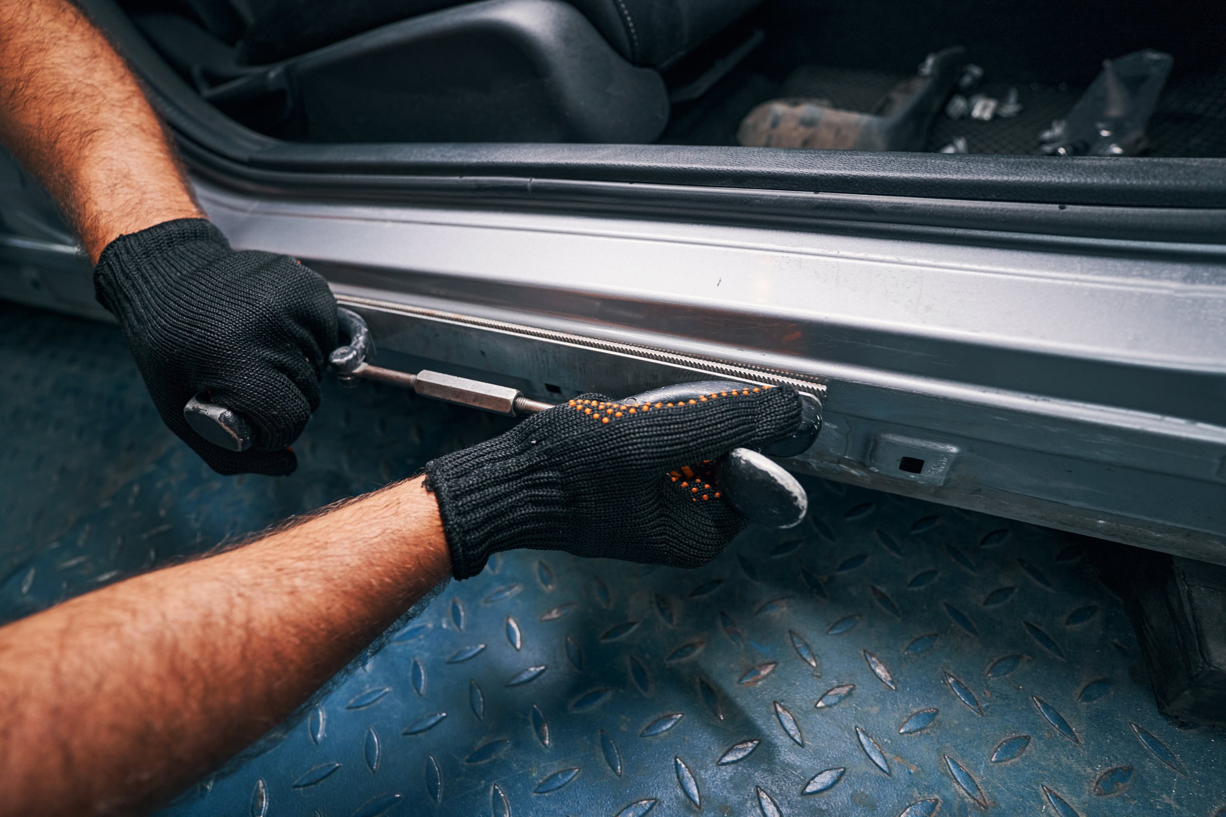 A person wearing black gloves using a wrench to work on the track of a vehicle, with the car door open and tools visible in the background.