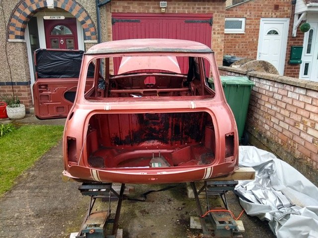 Vintage pink car body shell raised on jack stands in a driveway with the hood and back hatch removed, surrounded by tools and building elements.