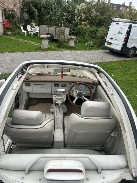 Looking into the interior of an old white convertible car parked outdoors, with visible seats, dashboard, and steering wheel, surrounded by a grassy area, trees, and nearby parked vehicles.