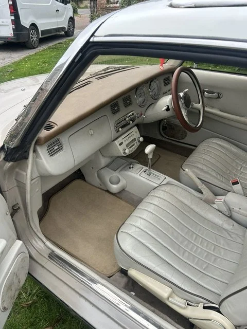 Interior of a vintage car showing a beige dashboard, steering wheel with a wood-like finish, manual gear shift, and gray upholstered seats.